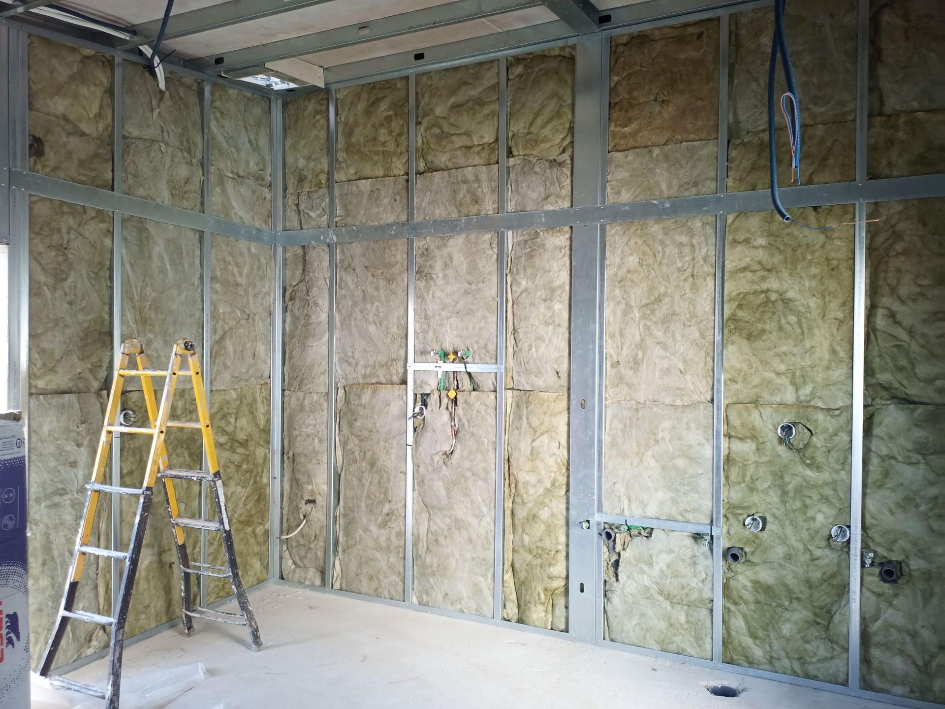 Parede interna em construção com estrutura metálica, isolamento e escada amarela.