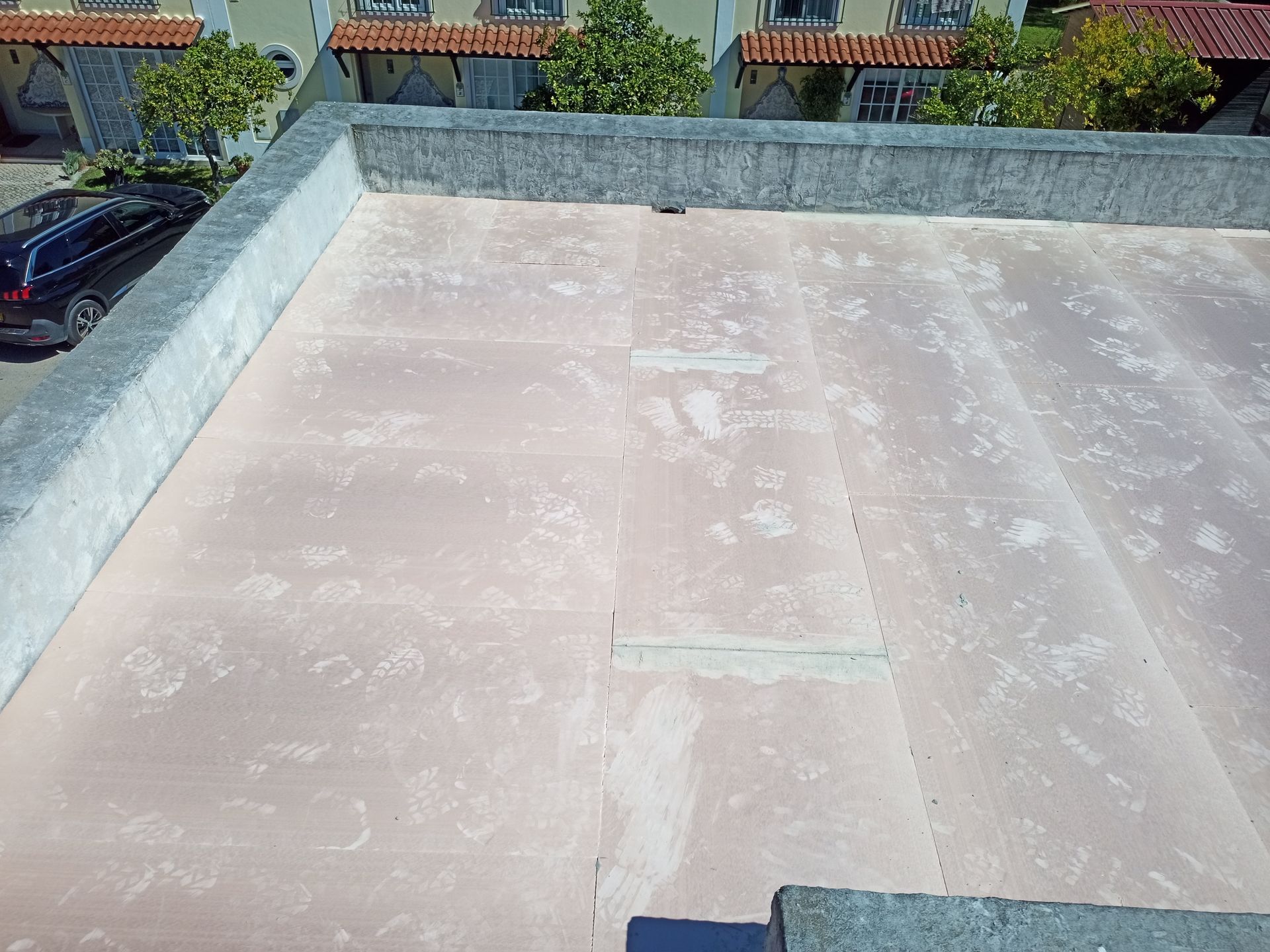 Telhado plano e claro com borda de concreto. Prédios residenciais e um carro são visíveis ao fundo.