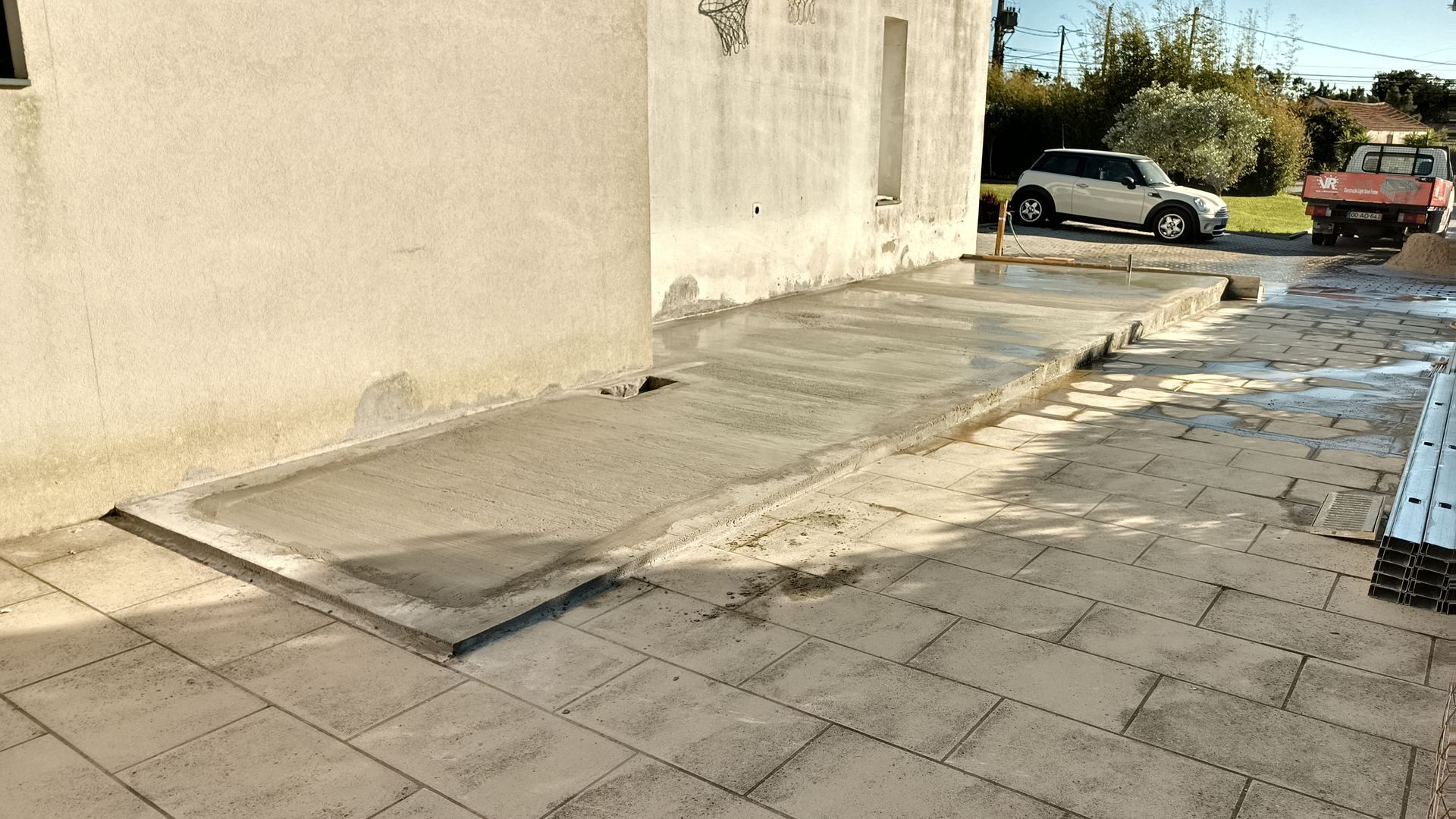 Rampa e pátio de concreto recém-construídos adjacentes a um edifício, com um veículo visível ao fundo.