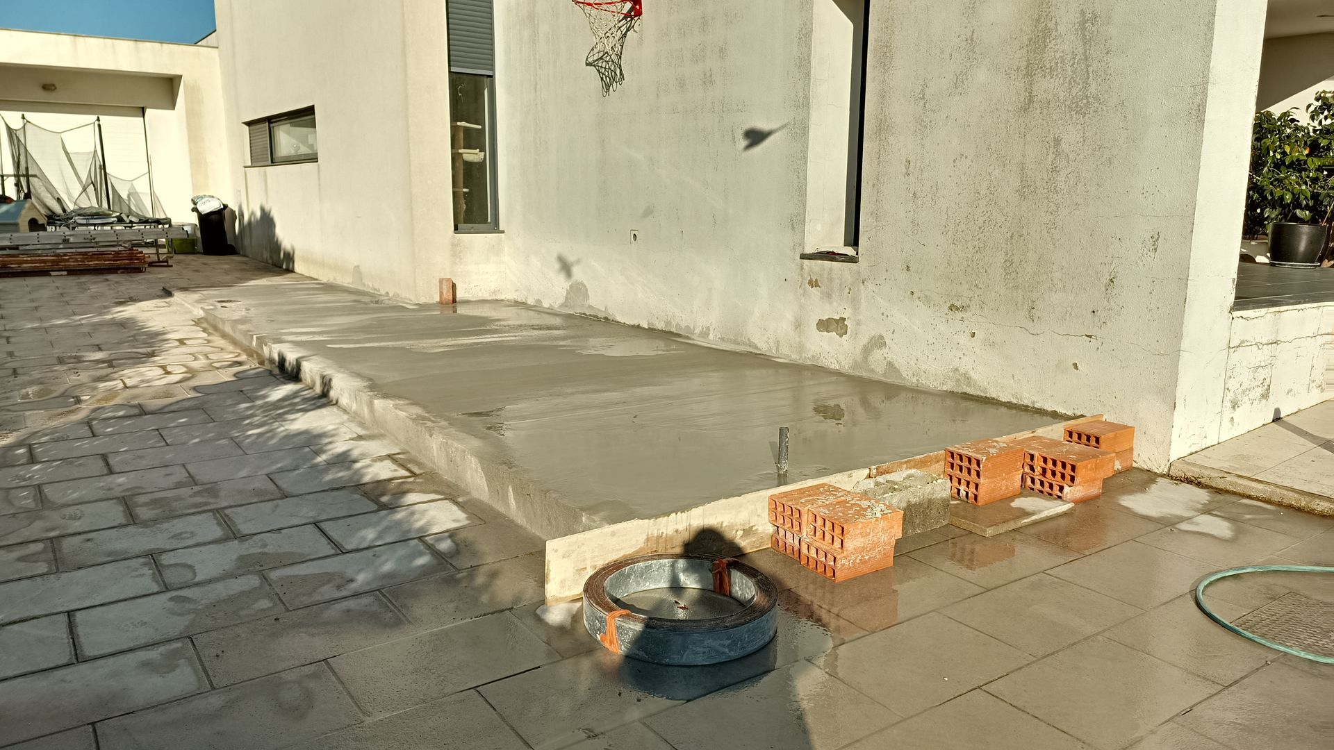 Um pátio de concreto sendo construído com tijolos, um balde e ferramentas ao lado de um prédio.