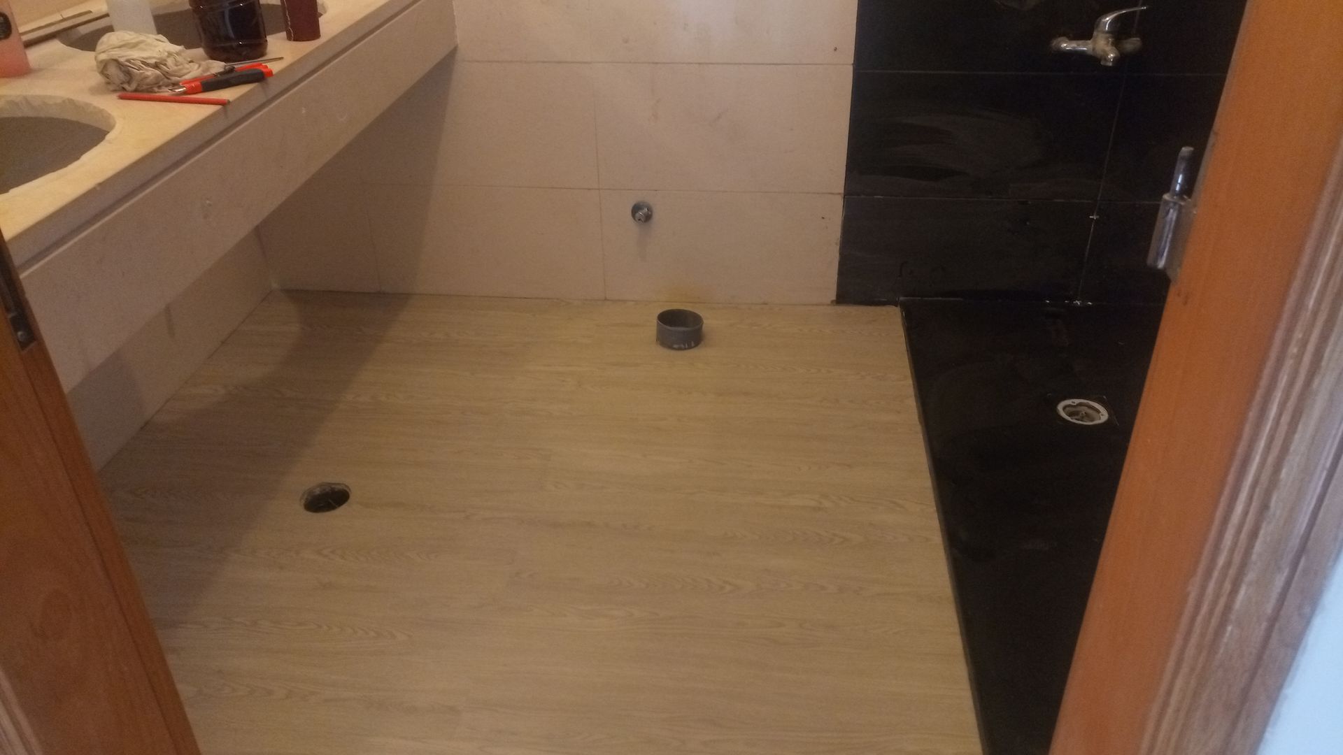 Interior de banheiro com piso de porcelanato imitando madeira clara. Pia e armário à esquerda. Área do chuveiro com azulejos escuros à direita.