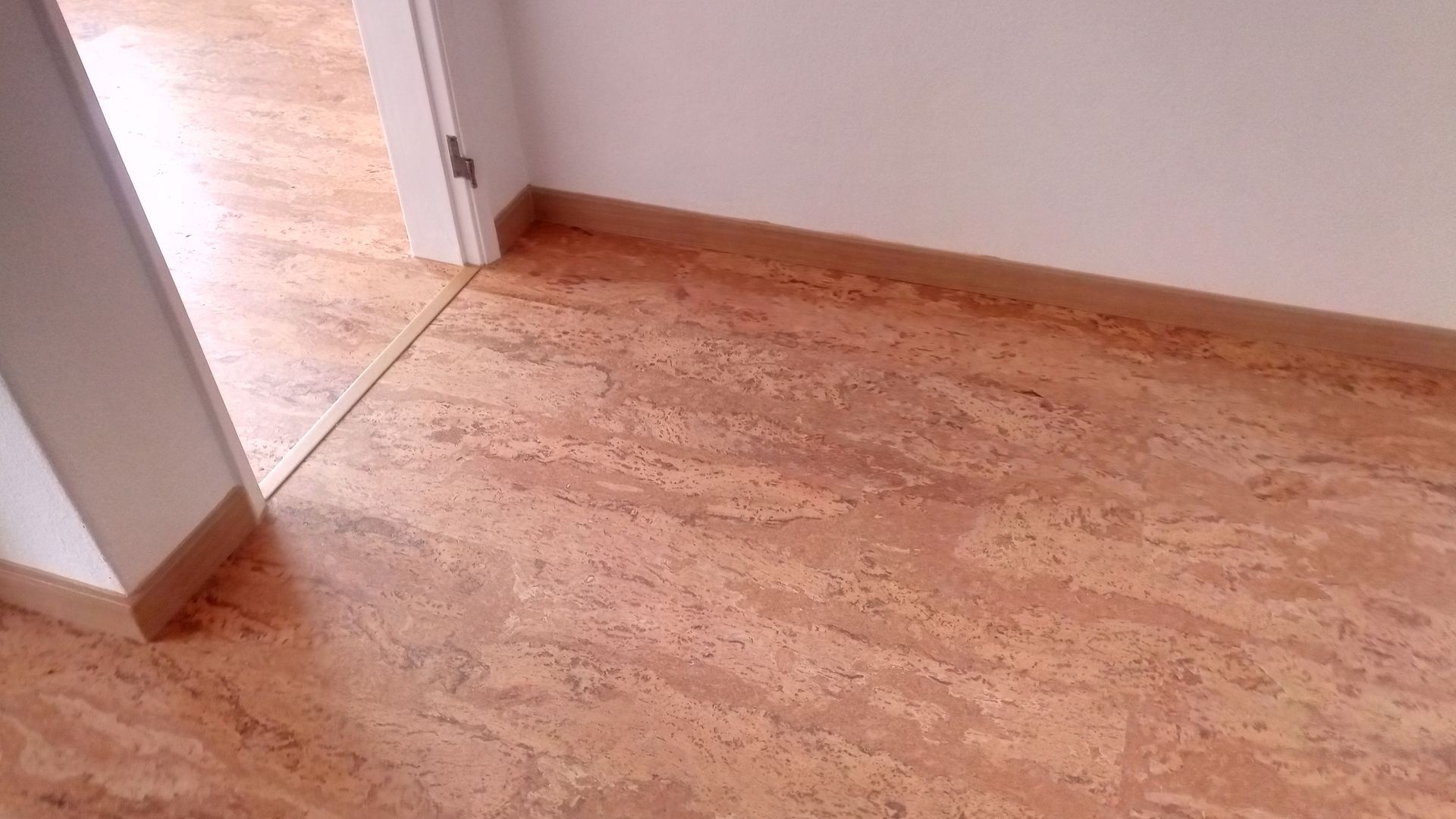 Piso de cortiça com veios castanho-claros, rodapés e porta de entrada.