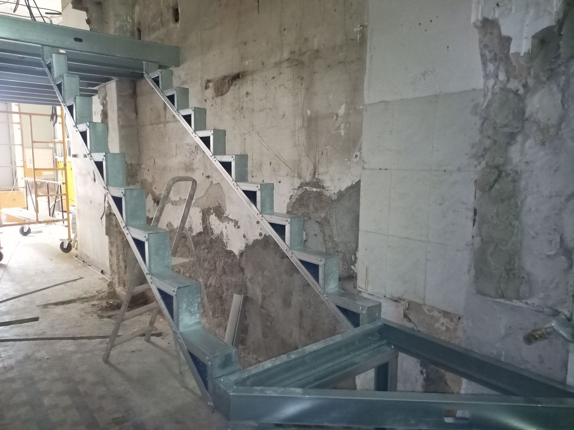 Estrutura metálica de escada em construção contra uma parede deteriorada, em ambiente interno.