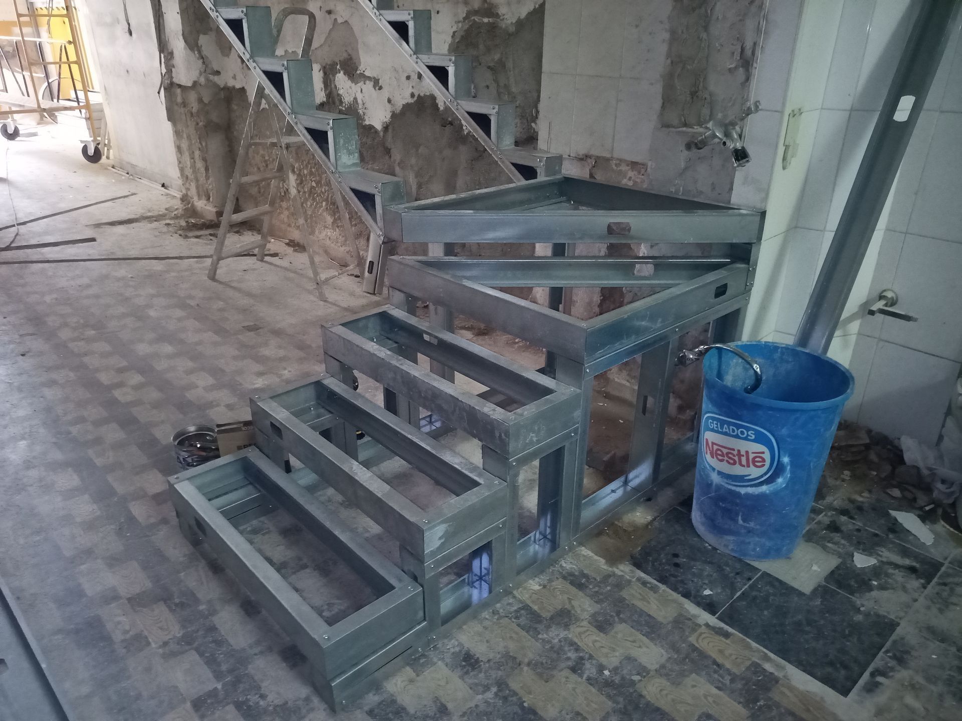 Estrutura metálica de escada em construção em uma sala com paredes descascando e um balde azul.
