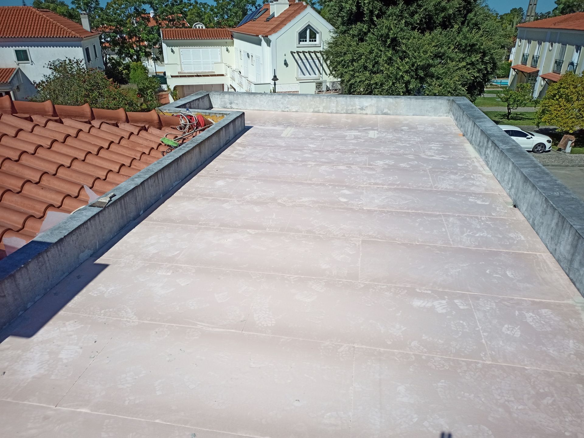 Telhado plano, superfície rosa claro, borda de concreto. Casas e árvores ao fundo.
