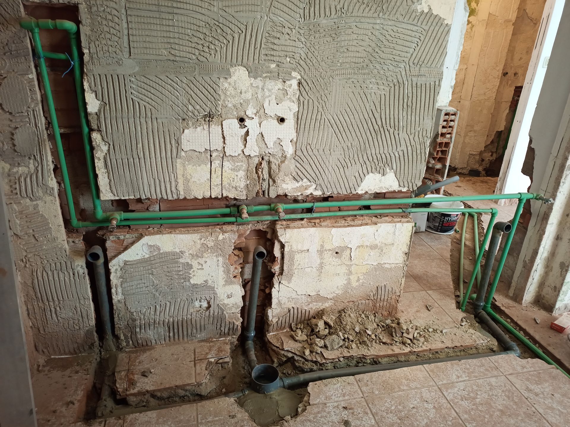 Obra de encanamento em andamento: canos verdes, parede exposta, canos de esgoto cinza.