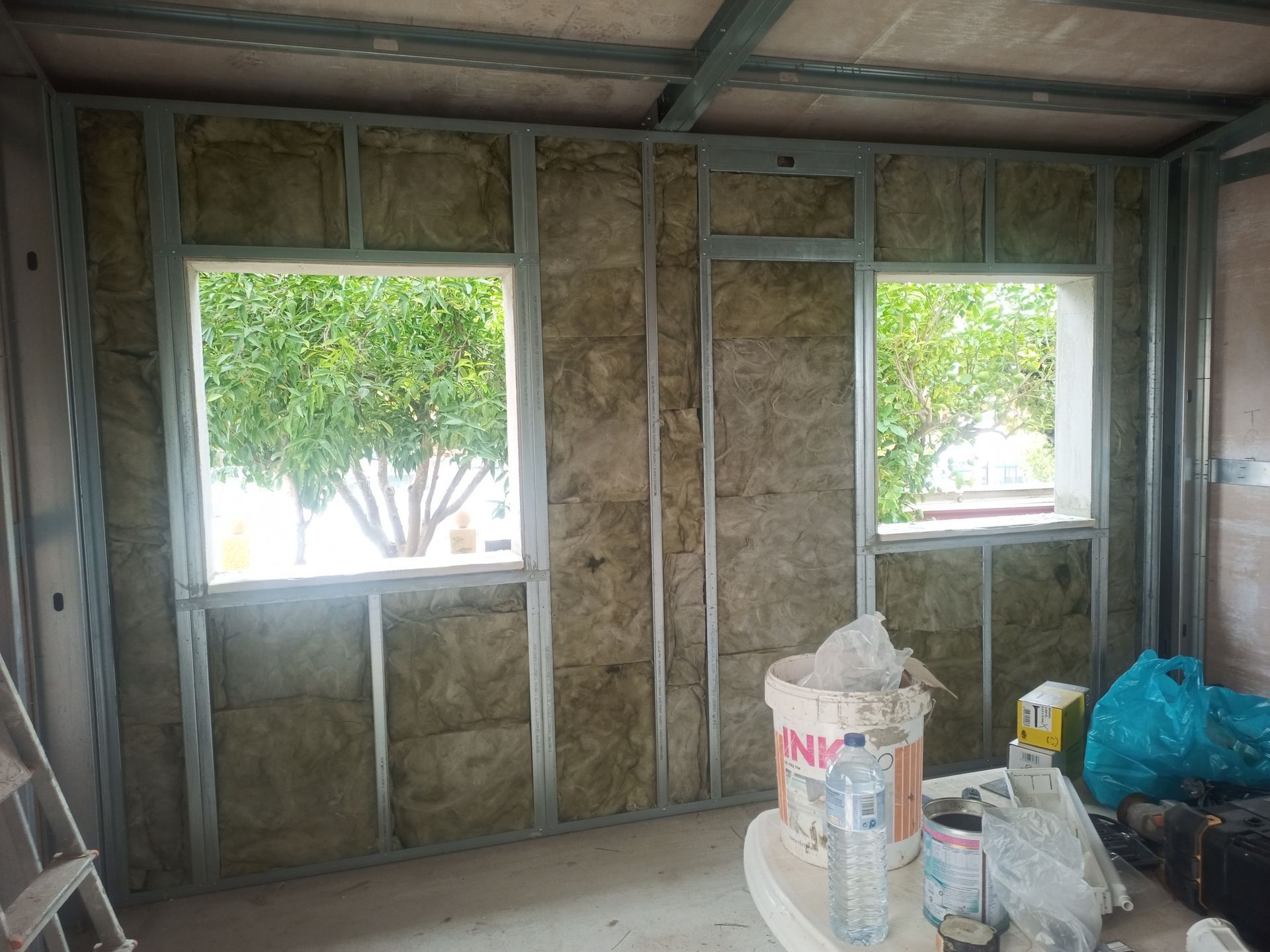 Vista interna de um cômodo em construção com estrutura metálica, isolamento e duas janelas.