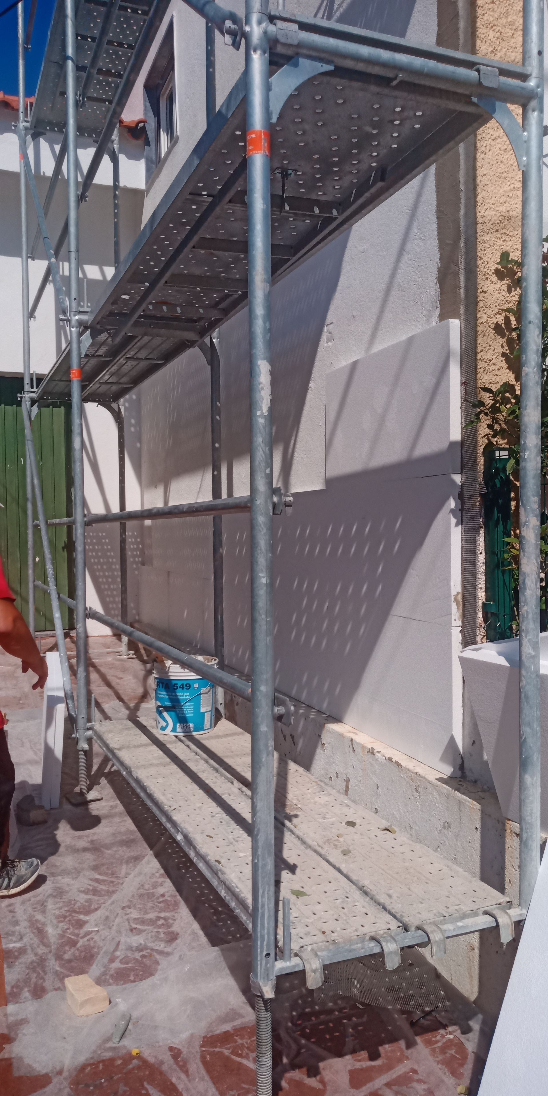 Andaimes erguidos contra a parede branca de um prédio. Um balde azul está no chão.