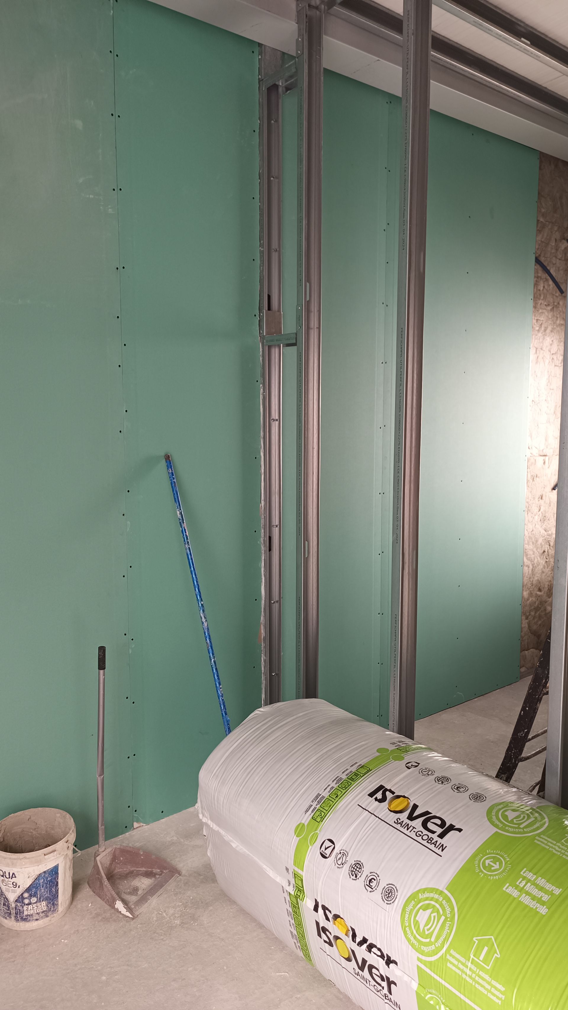 Cena de construção interna: drywall verde, estrutura metálica, rolo de isolamento, balde.
