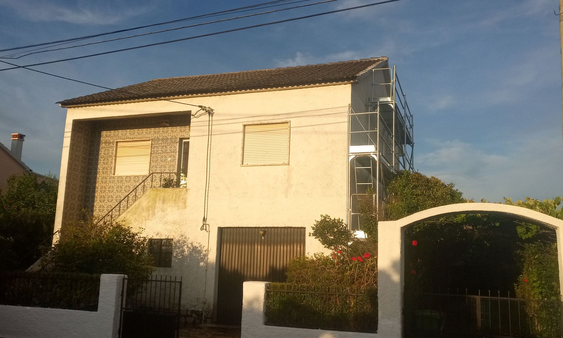 Casa branca de dois andares com porta de madeira fechada e janelas com venezianas. Andaimes na lateral.