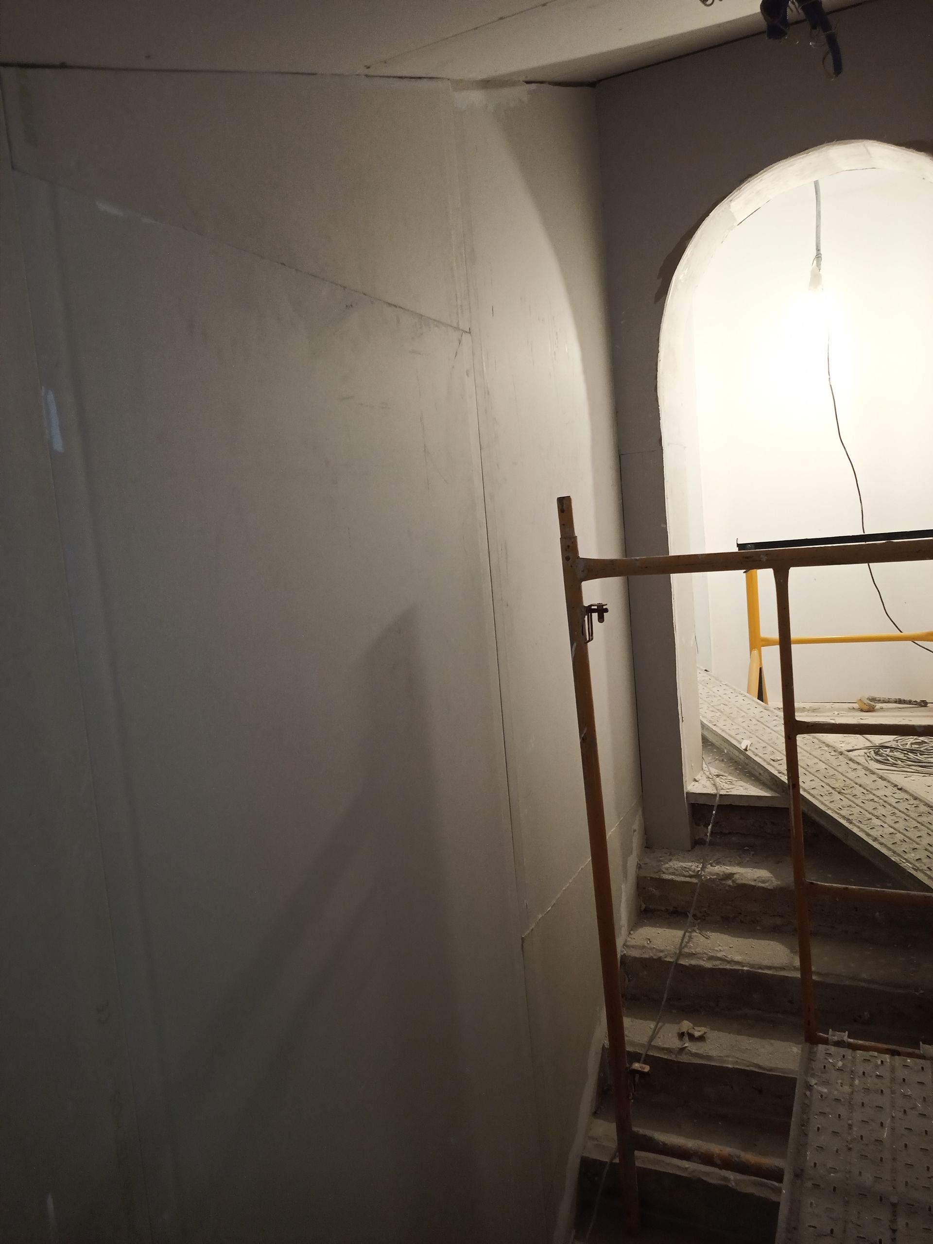 Cena de construção interna com paredes inacabadas, porta em arco e andaimes na escada.