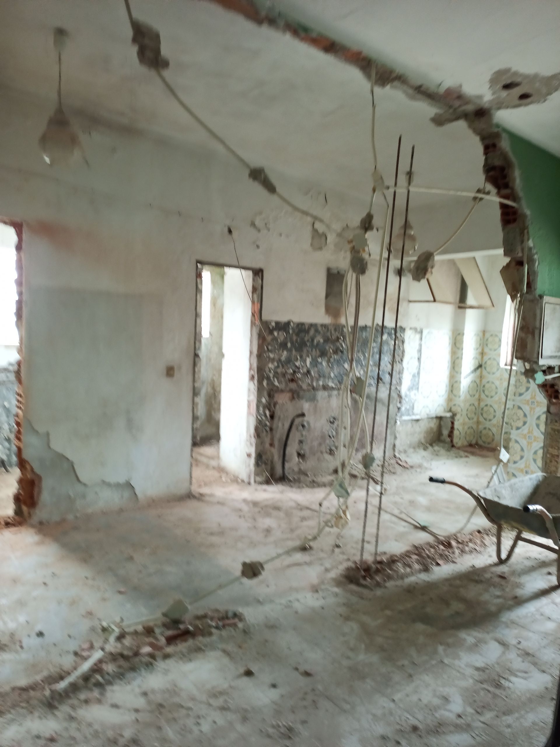 Demolição interna de um edifício. Paredes e teto parcialmente removidos. Fiação elétrica exposta. Carrinho de mão presente.