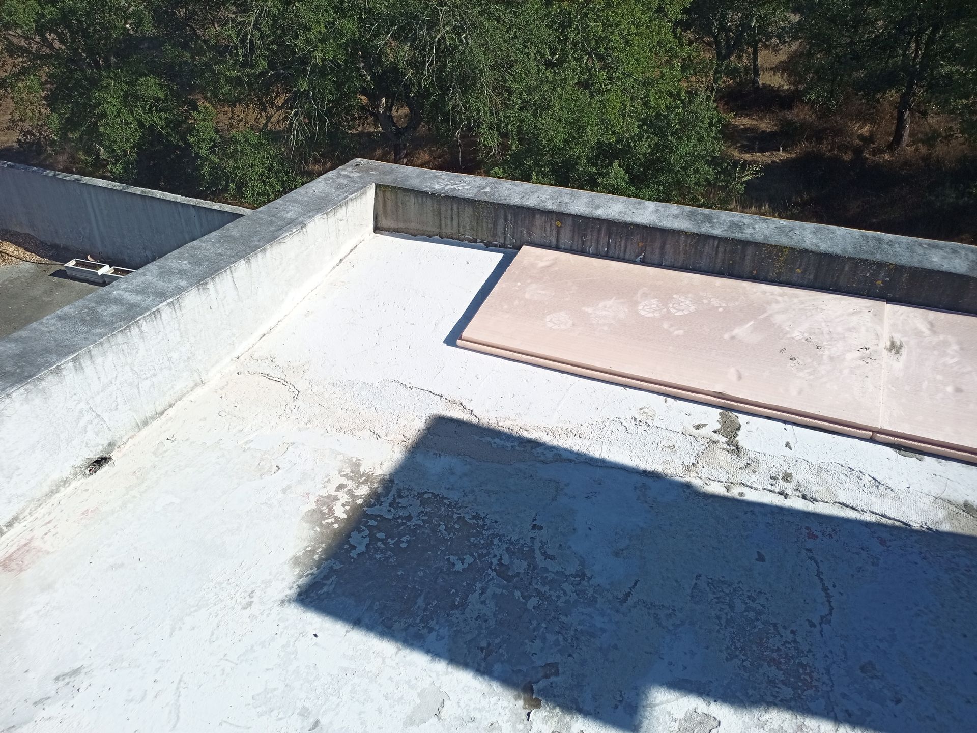 Um terraço plano com bordas de concreto, um painel marrom claro e árvores ao fundo sob um céu ensolarado.