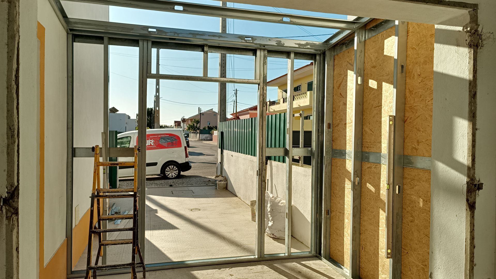 Vista interna de um edifício em construção, mostrando a estrutura metálica de uma janela com vista para uma rua com veículos estacionados.