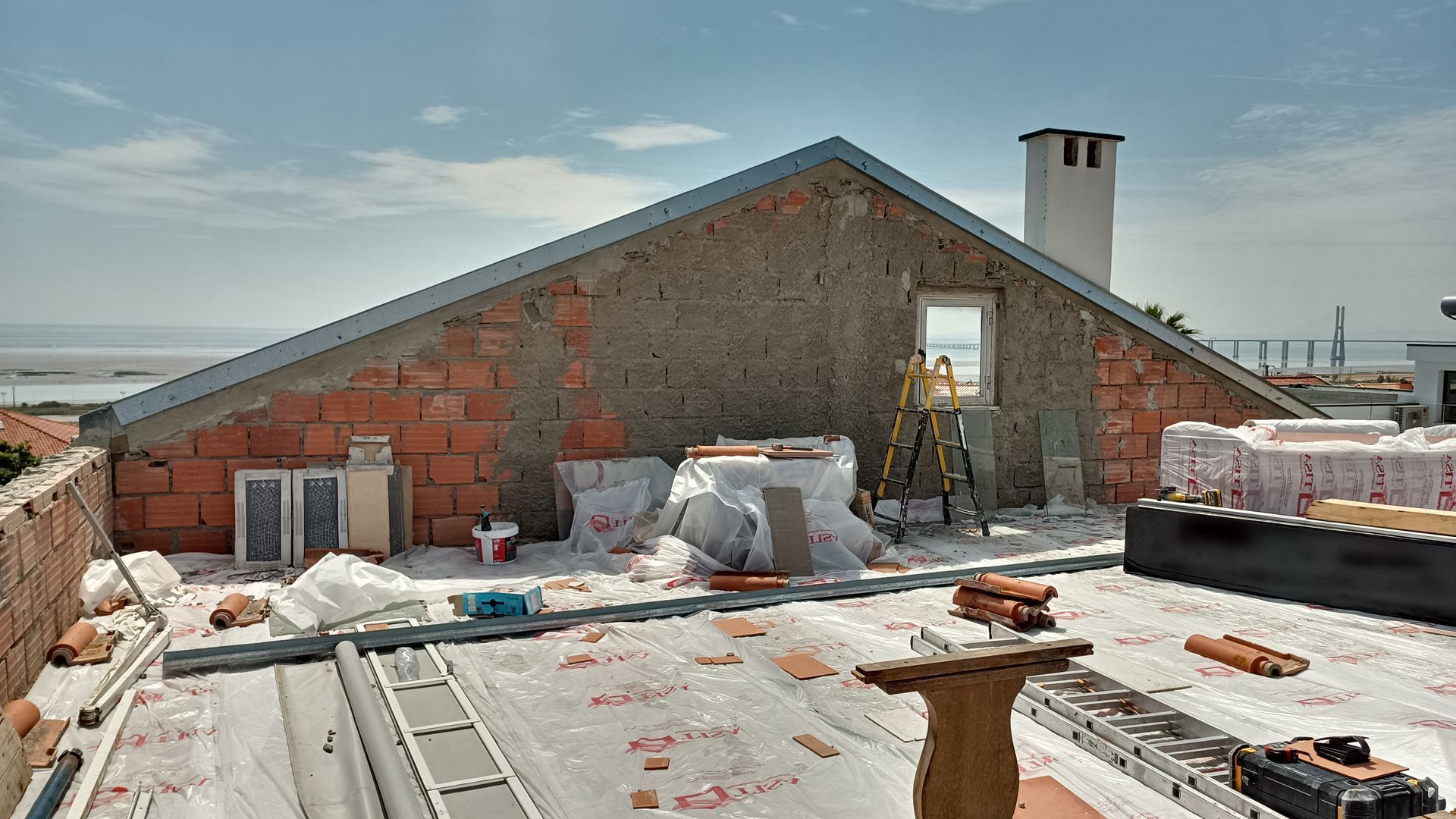 Canteiro de obras em um telhado com parede de tijolos, materiais e uma escada sob um céu azul.