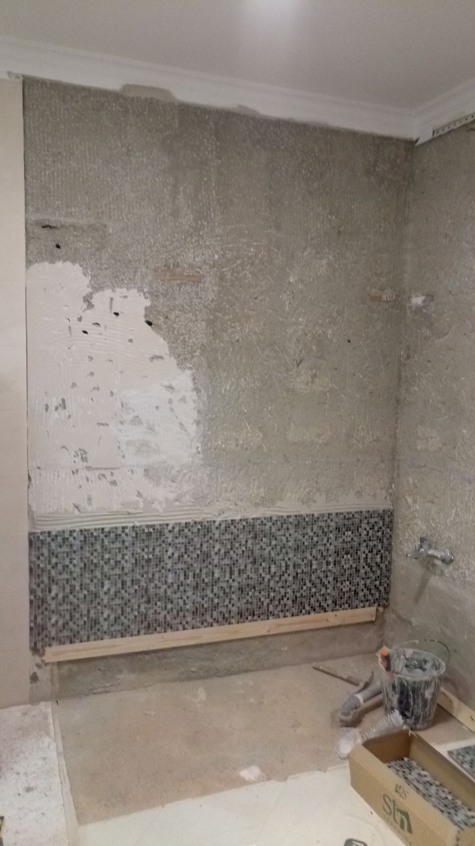 Parede do banheiro em construção, parcialmente revestida com azulejos. Gesso cinza, azulejos de mosaico, madeira e ferramentas visíveis.