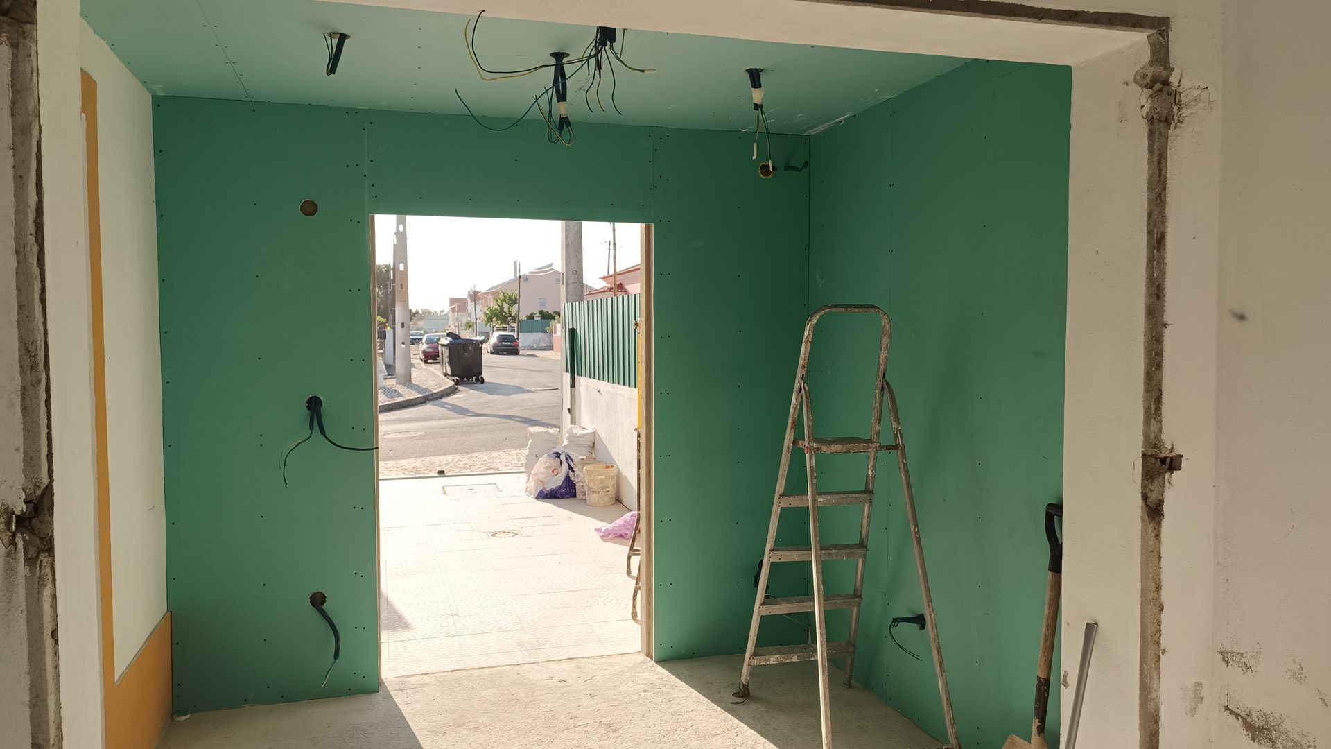 Quarto em construção com paredes verdes, porta, escada e fiação exposta. Vista para a rua.