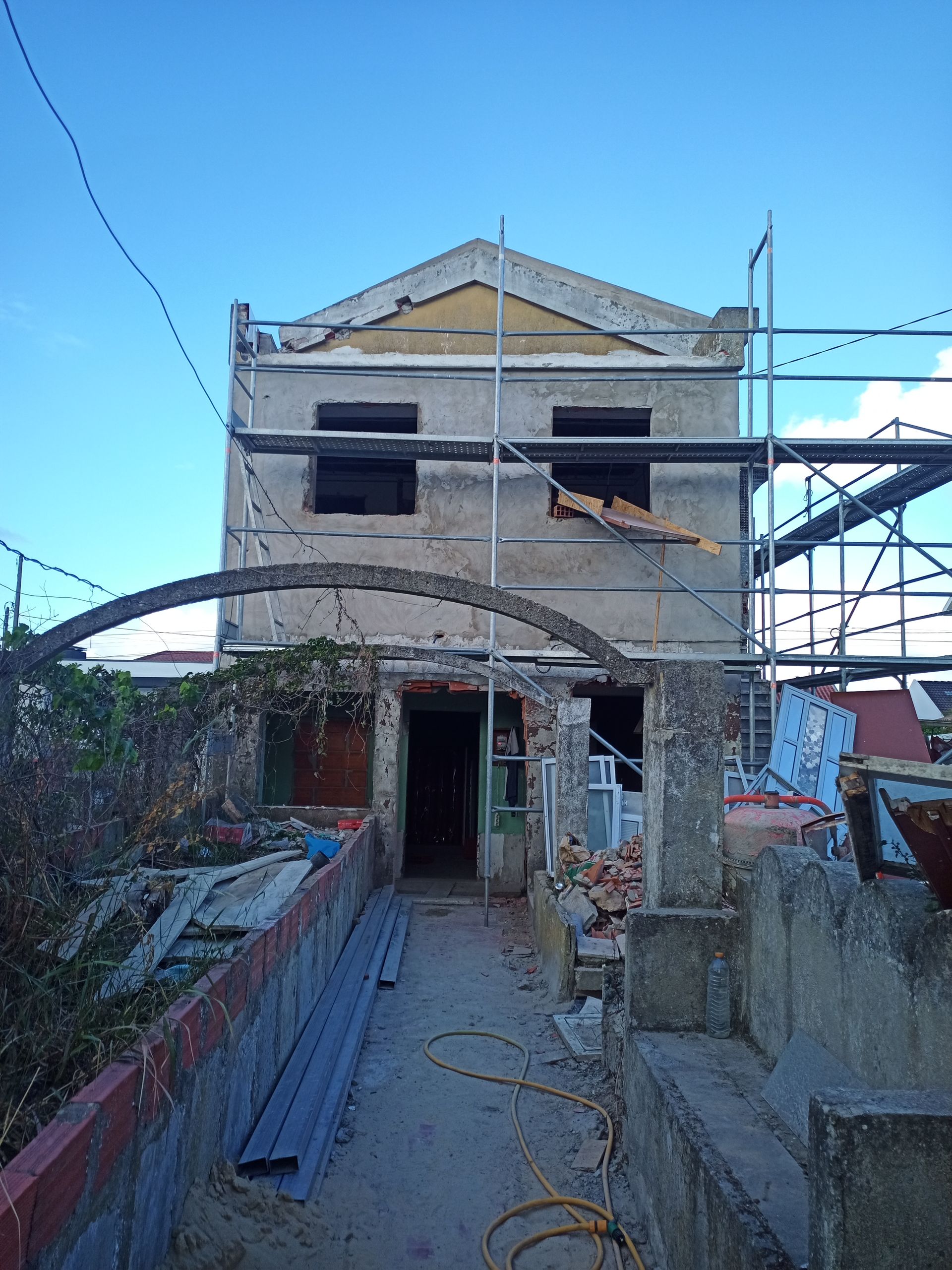 Prédio de dois andares em reforma, com andaimes e entrada em arco; céu azul ao fundo.