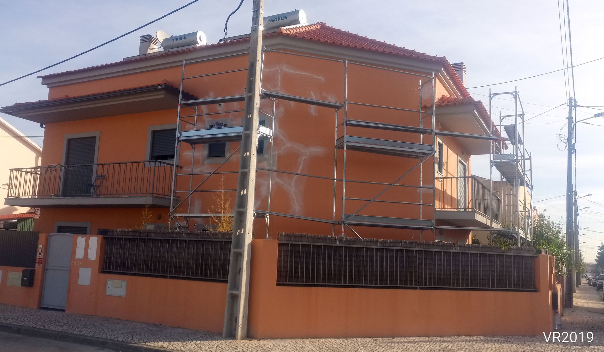 Casa laranja de dois andares com andaimes para reformas. Cerca marrom e telhado de telha vermelha.