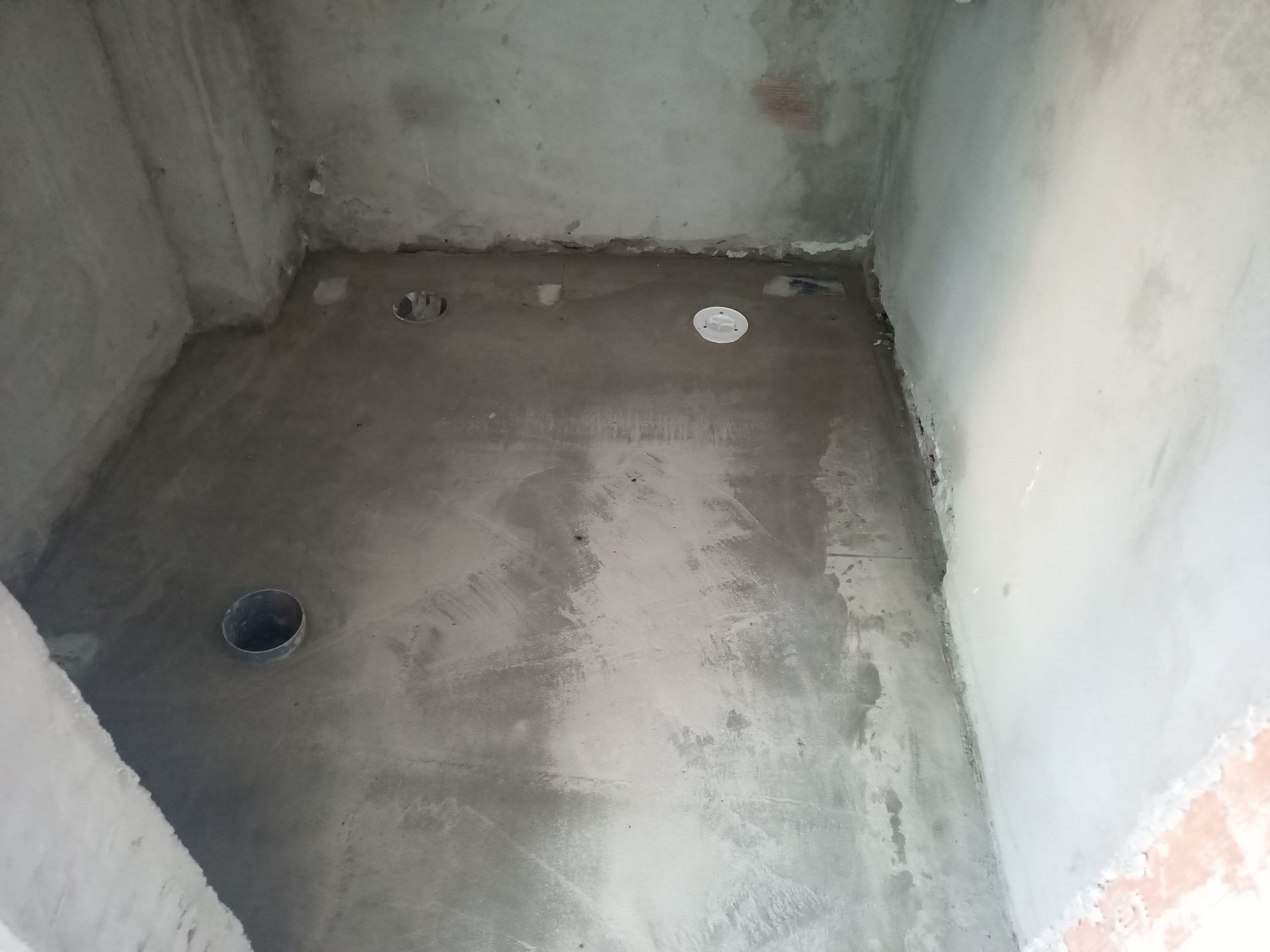 Quarto inacabado com paredes de concreto, ralos no piso e instalações hidráulicas no piso e nas paredes.