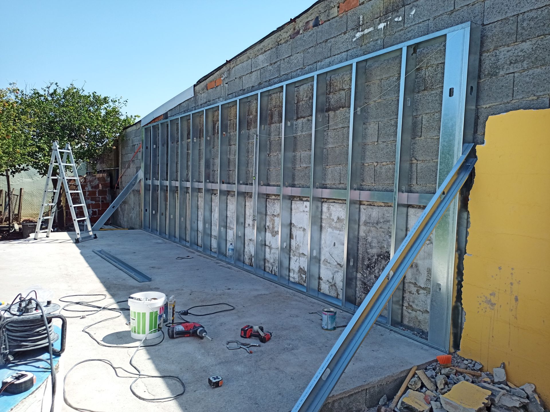 Estrutura metálica instalada em uma parede de blocos de concreto, sustentada por escoras temporárias. Canteiro de obras, área externa, ensolarada.