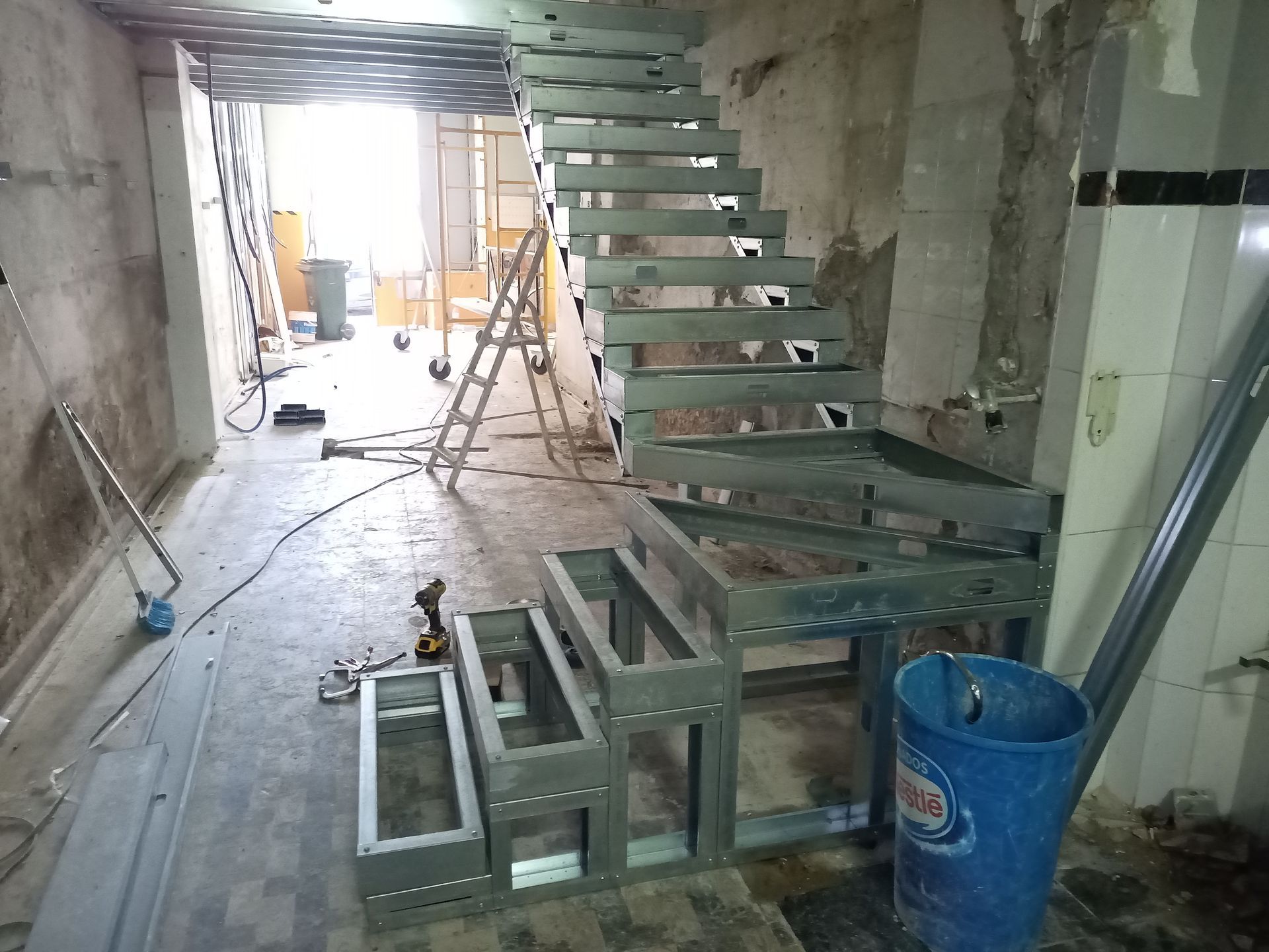 Escada metálica em construção em um cômodo com paredes e piso inacabados, com ferramentas e uma escada presentes.