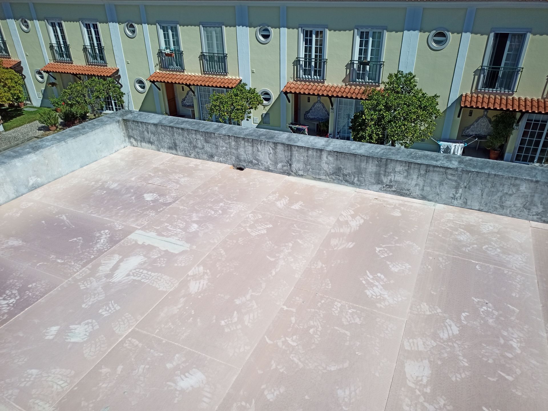 Vista de um terraço plano com uma barreira de concreto; um prédio com janelas e varandas está ao fundo.