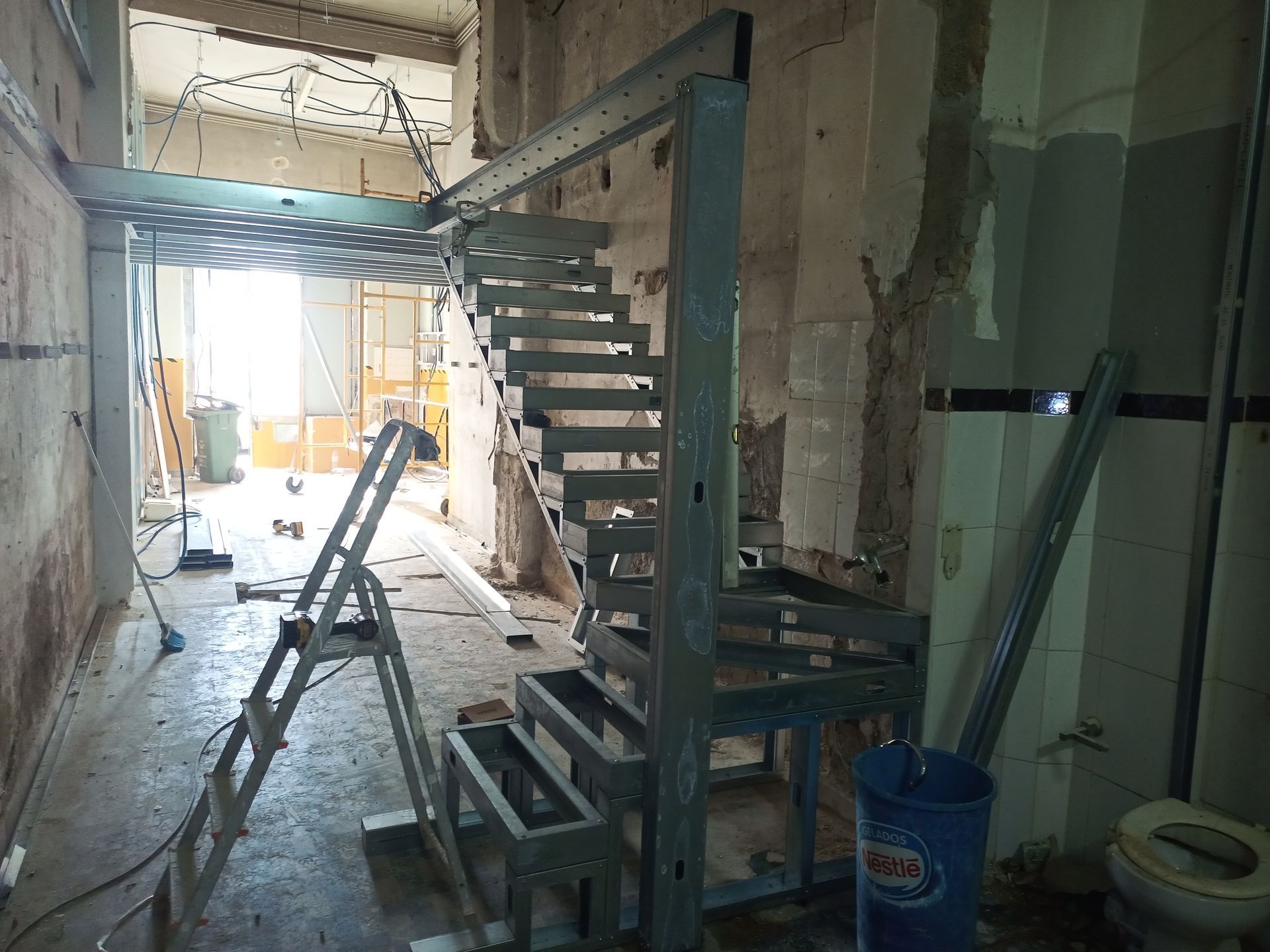 Escada metálica em construção em um cômodo inacabado. Uma plataforma metálica perfurada está localizada acima da escada.