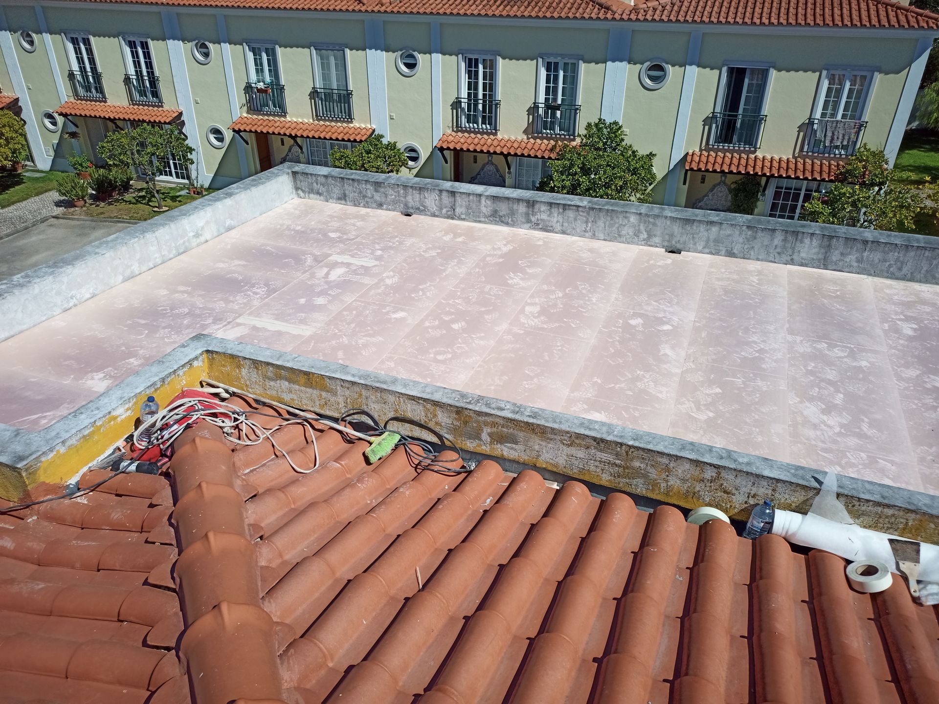 Telhado de telhas vermelhas em primeiro plano, com um telhado plano de cor clara e um edifício com varandas ao fundo.