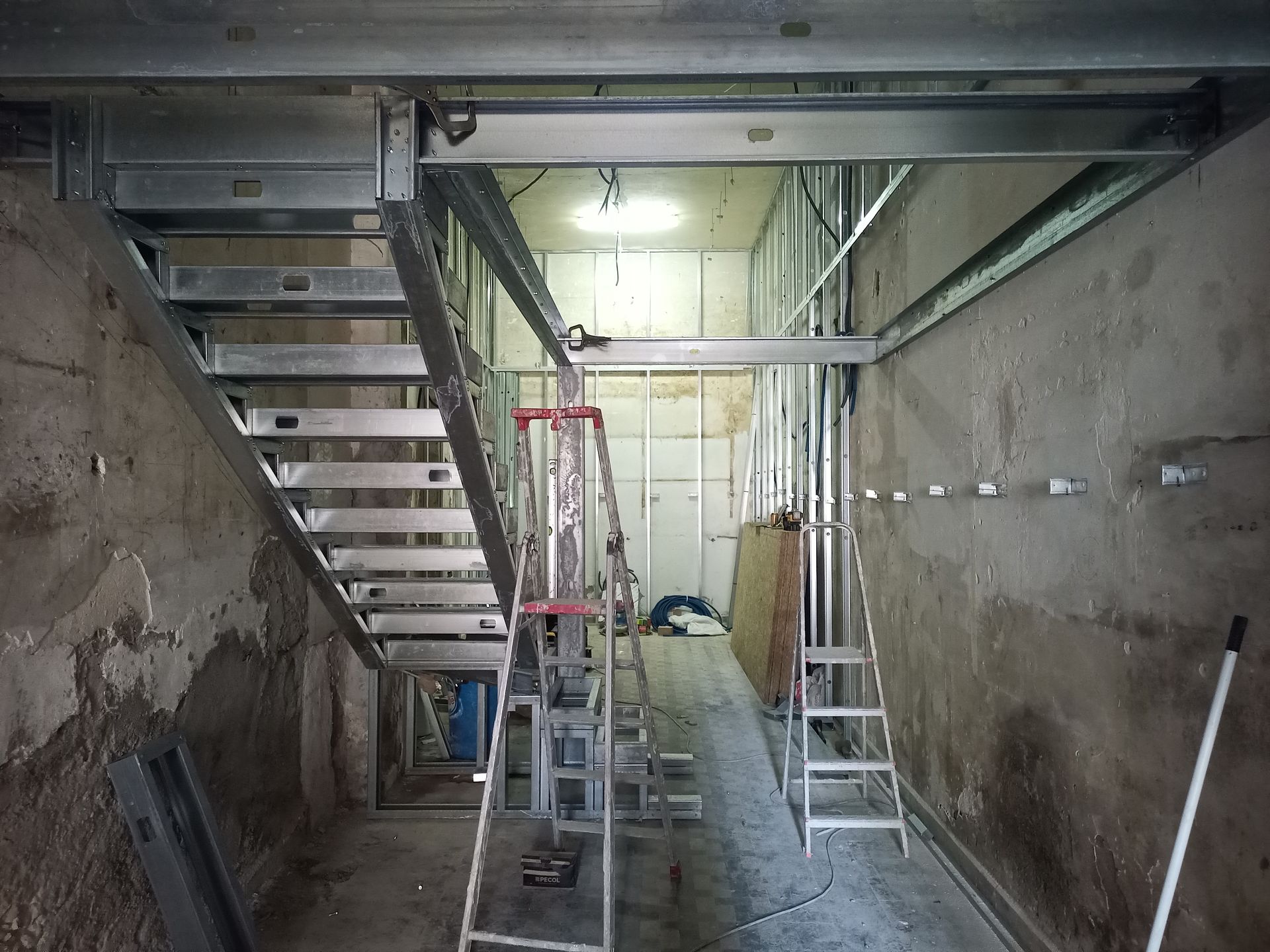 Canteiro de obras com escada e estrutura metálica. Presença de escada tipo cavalete. Paredes e teto de concreto.