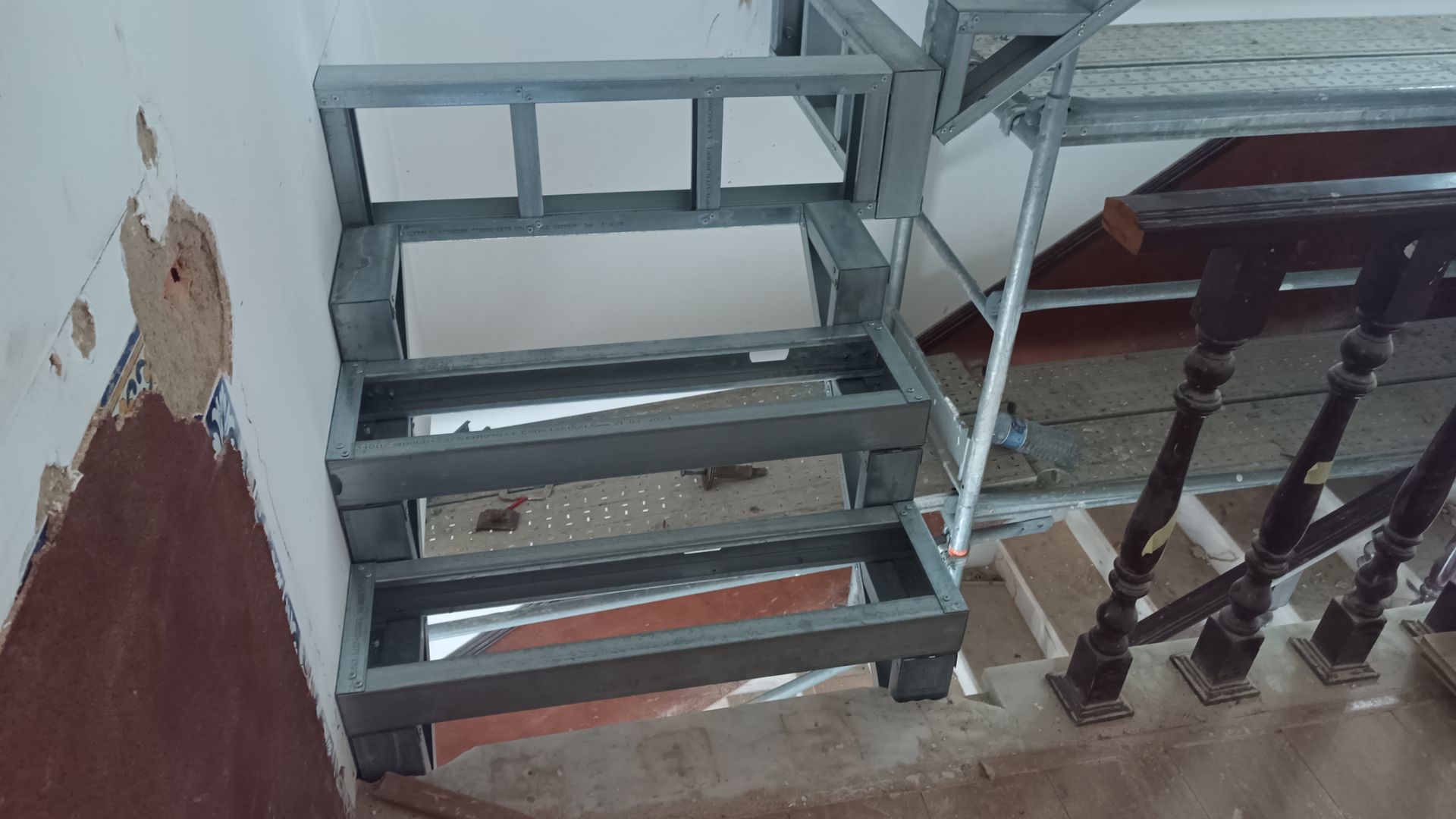 Estrutura metálica de uma escada sendo instalada perto de uma parede com reboco danificado.