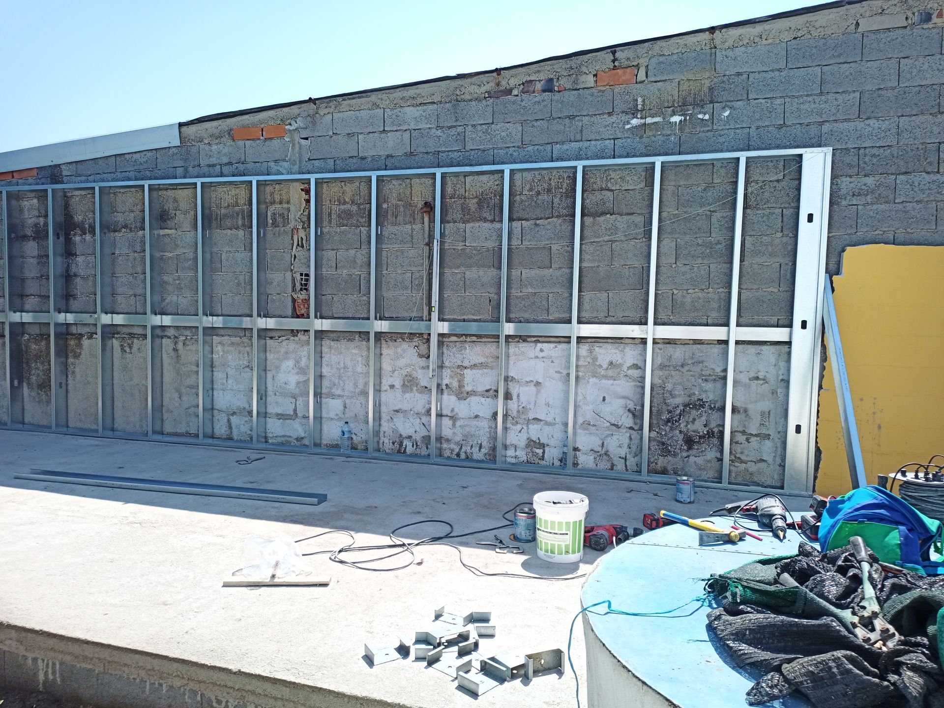 Construção de parede com estrutura metálica contra uma parede de blocos de concreto, com ferramentas e materiais no chão.