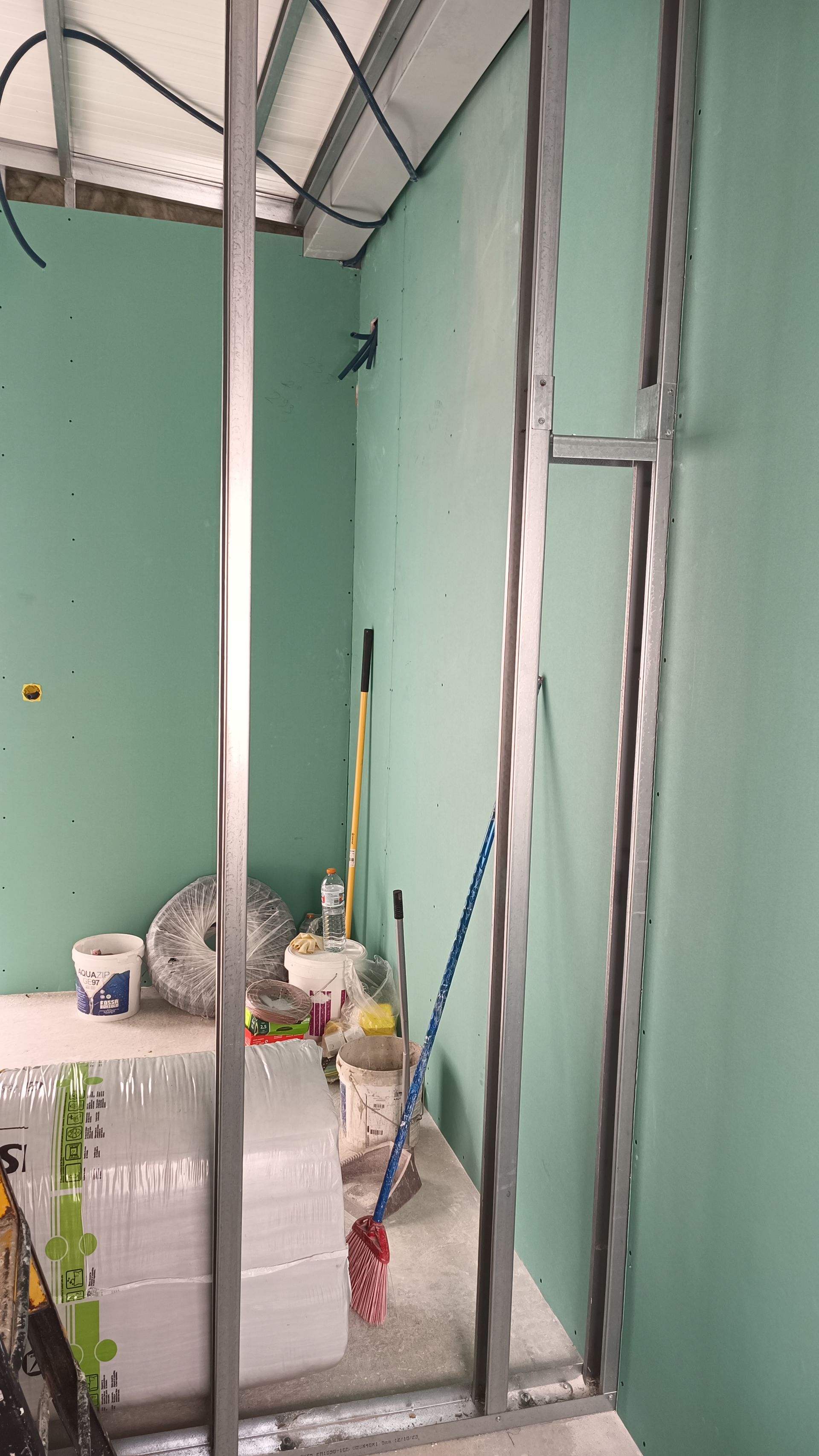 Interior de uma obra com estrutura metálica formando uma parede. É possível ver gesso acartonado verde e ferramentas.