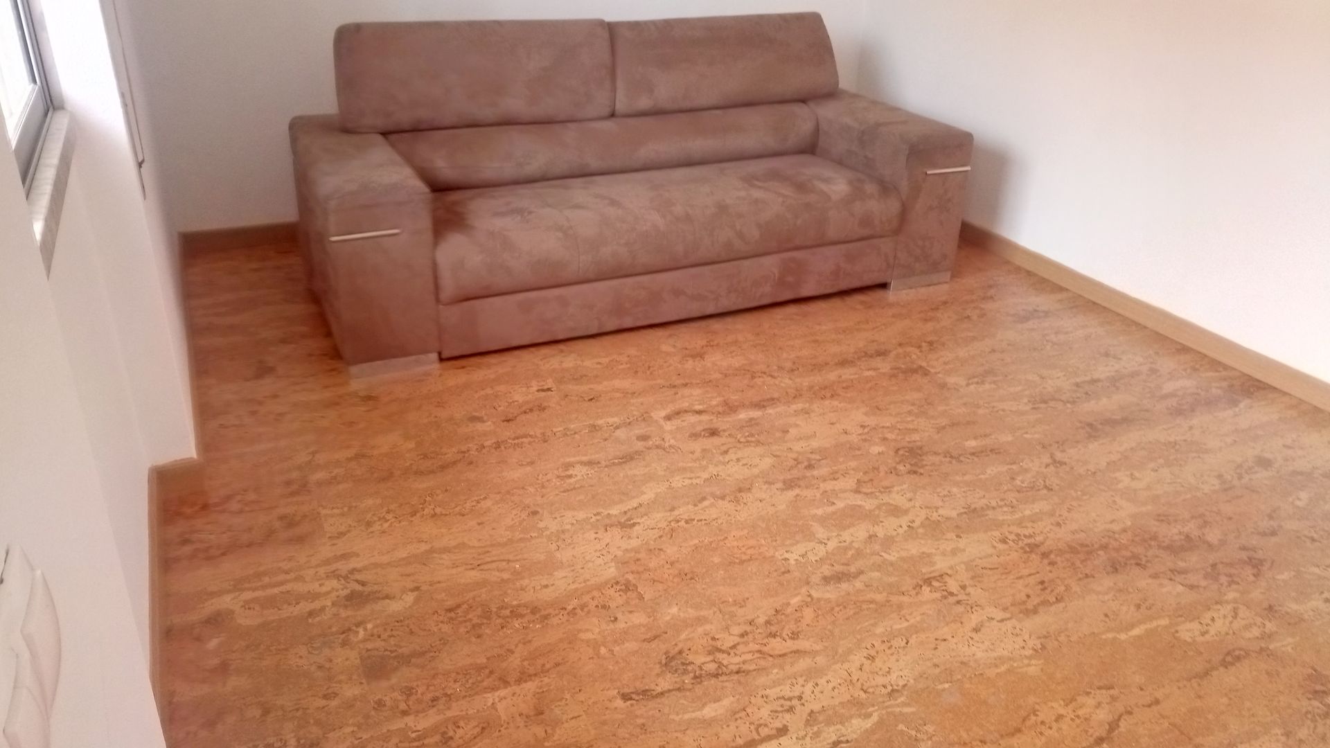 Sofá marrom sobre piso marrom em uma sala com paredes brancas e uma janela.