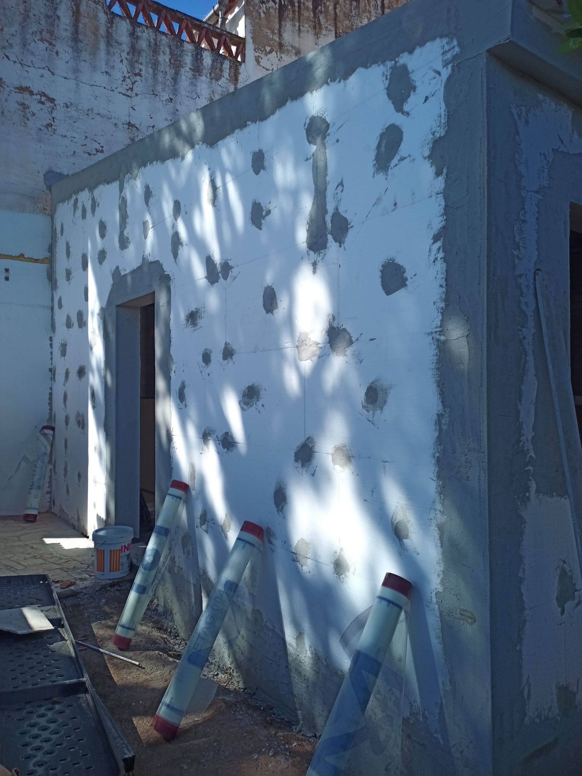 Parede externa com reboco branco e manchas de adesivo espalhadas, batente da porta e suportes. Canteiro de obras.