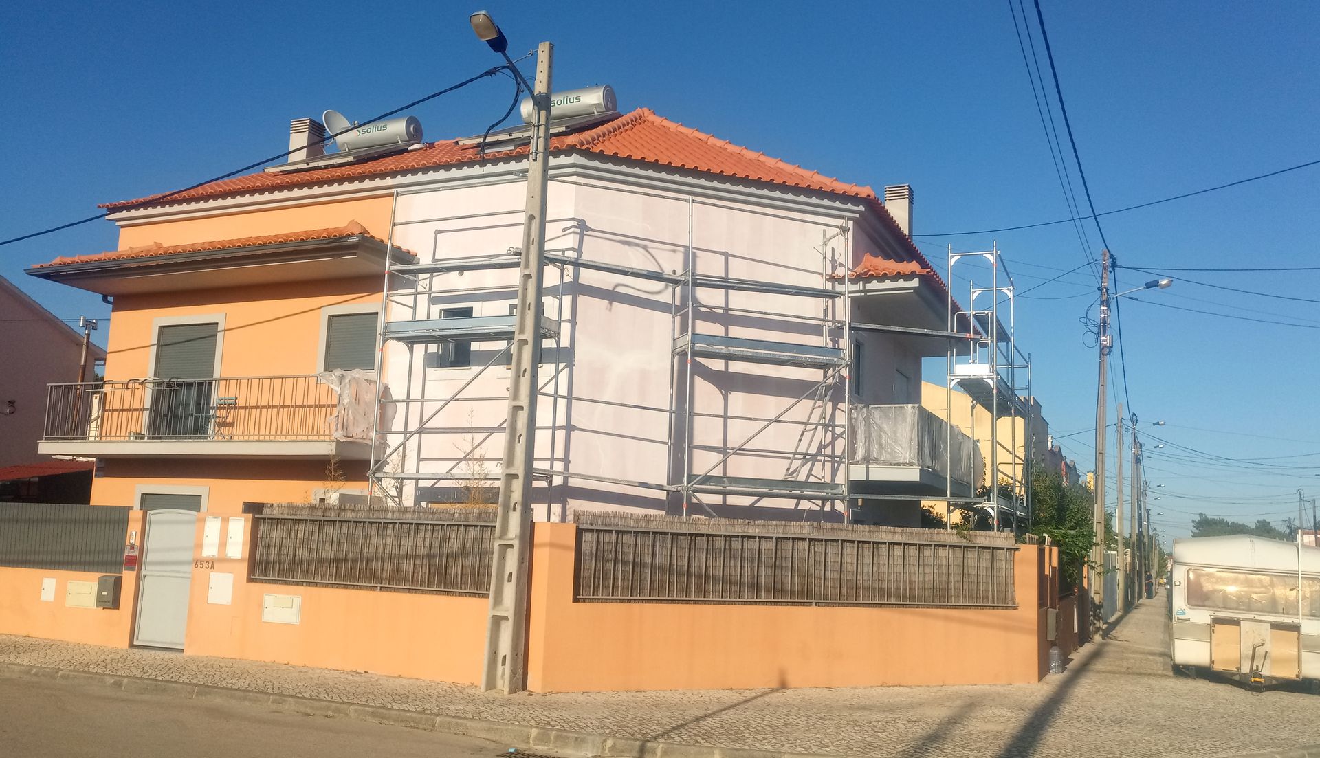 Casa em construção com andaimes, paredes laranja e rosa, painéis solares no telhado, vista da rua.