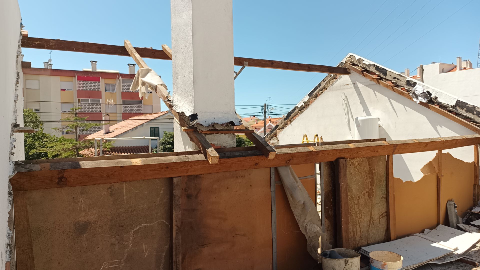 Canteiro de obras com vigas de madeira expostas, paredes antigas e parte do telhado. Prédios da cidade ao fundo.