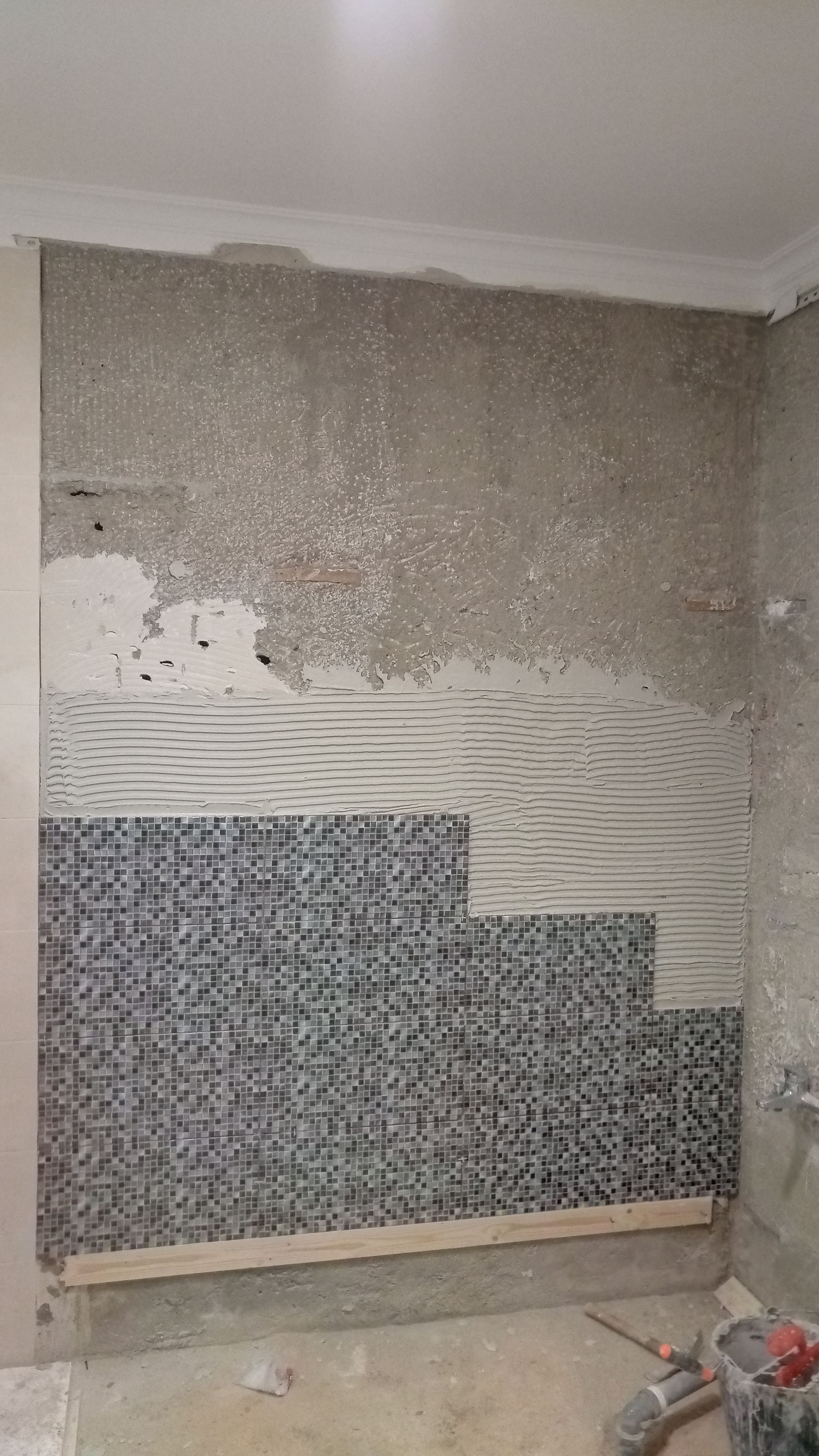 Parede com mosaico em construção, mostrando cimento, adesivo e uma base de madeira.