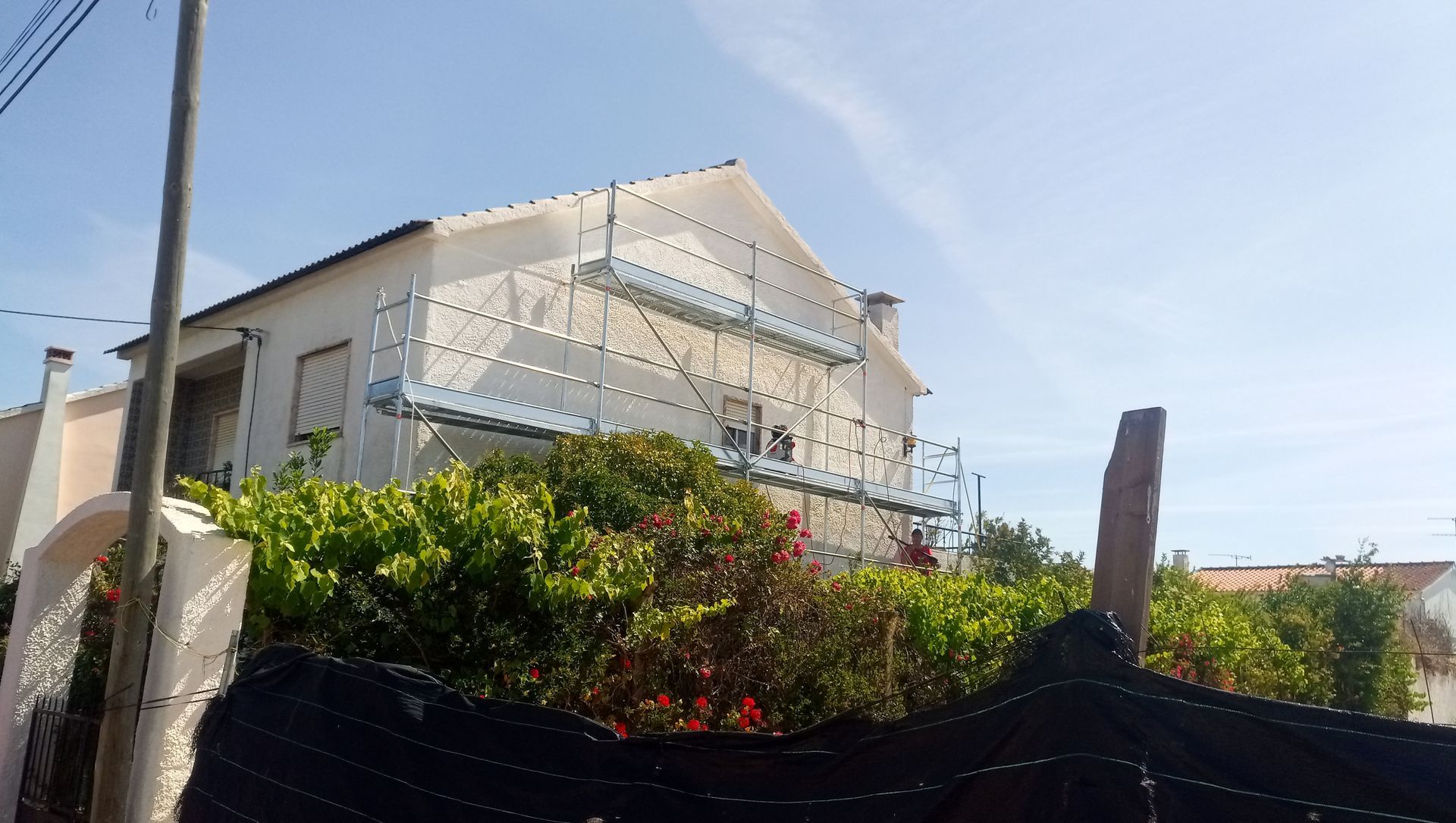 Uma casa com andaimes na parede, tendo como pano de fundo um céu azul. Arbustos verdejantes em primeiro plano.