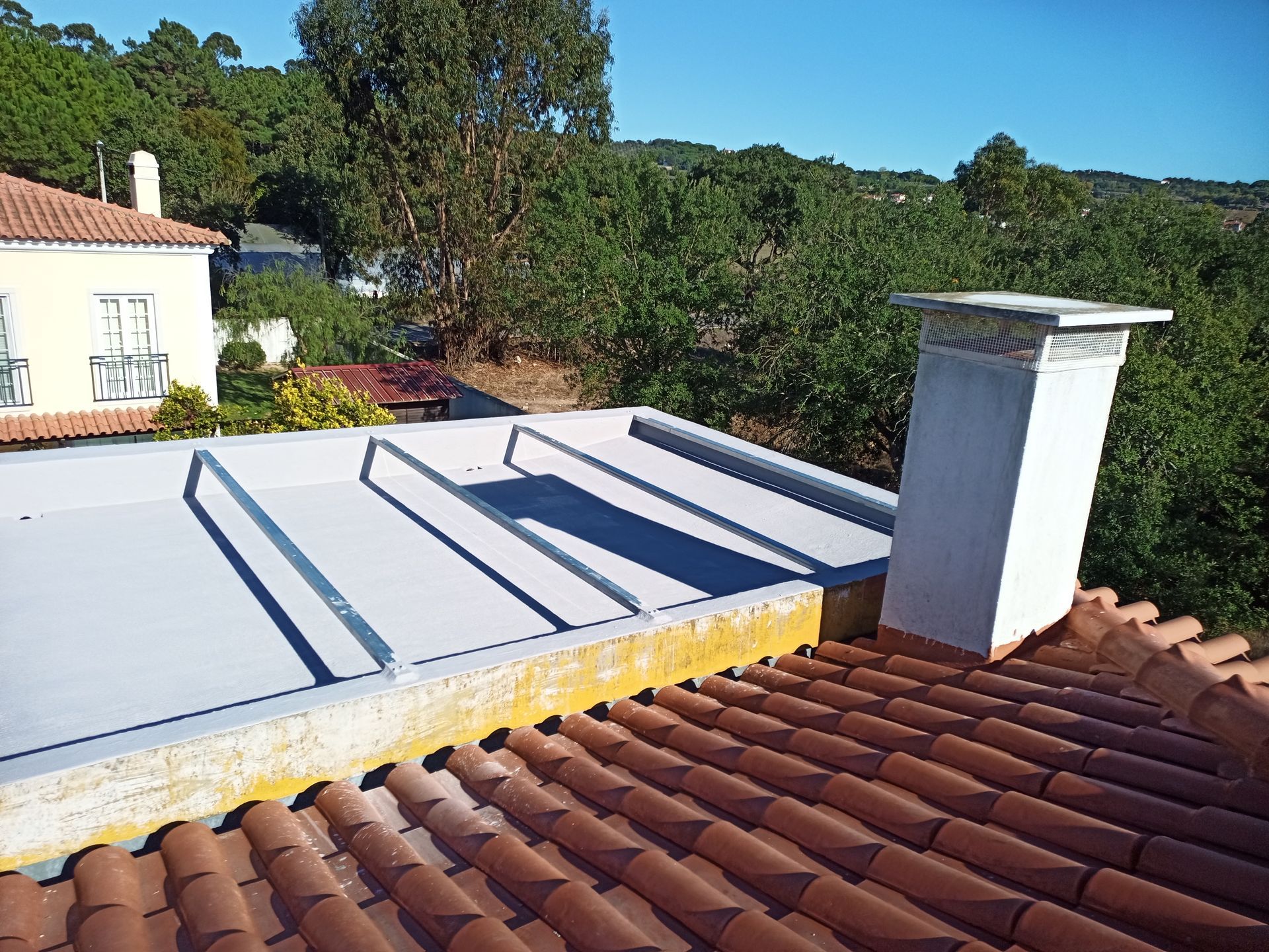 Vista do telhado de uma casa com telhas vermelhas, uma seção plana de telhado cinza e uma chaminé branca contra um fundo de árvores.