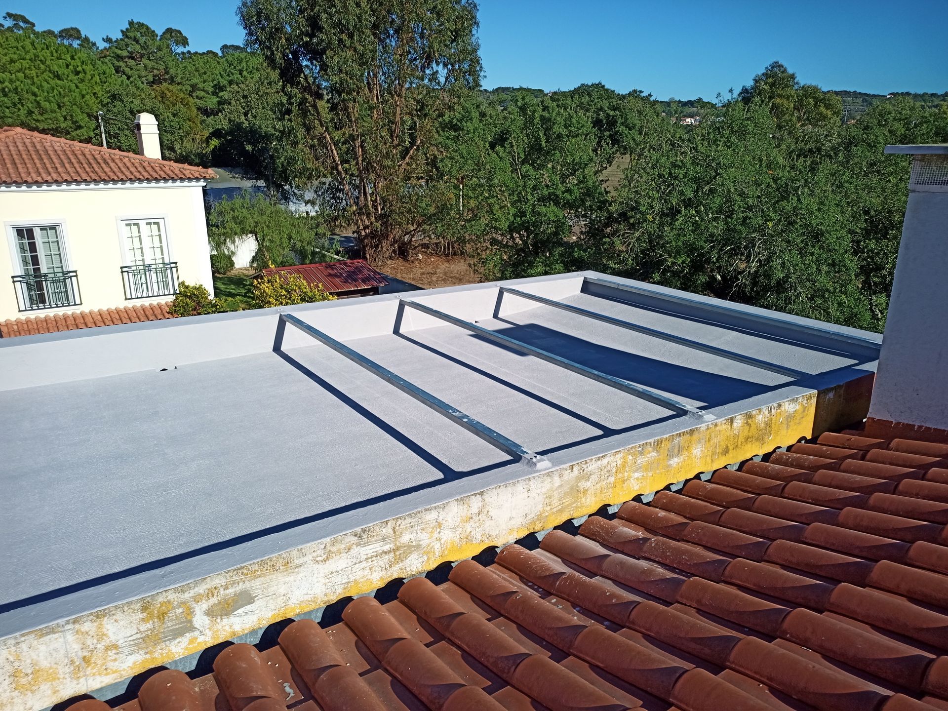 Telhado plano cinza com vigas de metal, delimitado por concreto amarelado, adjacente a um telhado de telhas vermelhas.