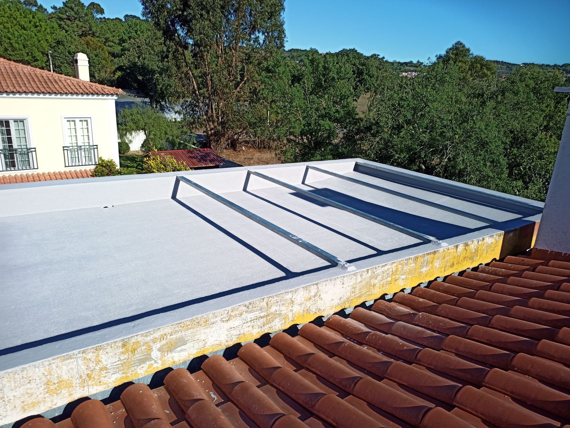 Telhado plano, cinza-claro, com bordas elevadas, visível a partir de um telhado mais baixo, coberto com telhas. Árvores e um prédio ao fundo.