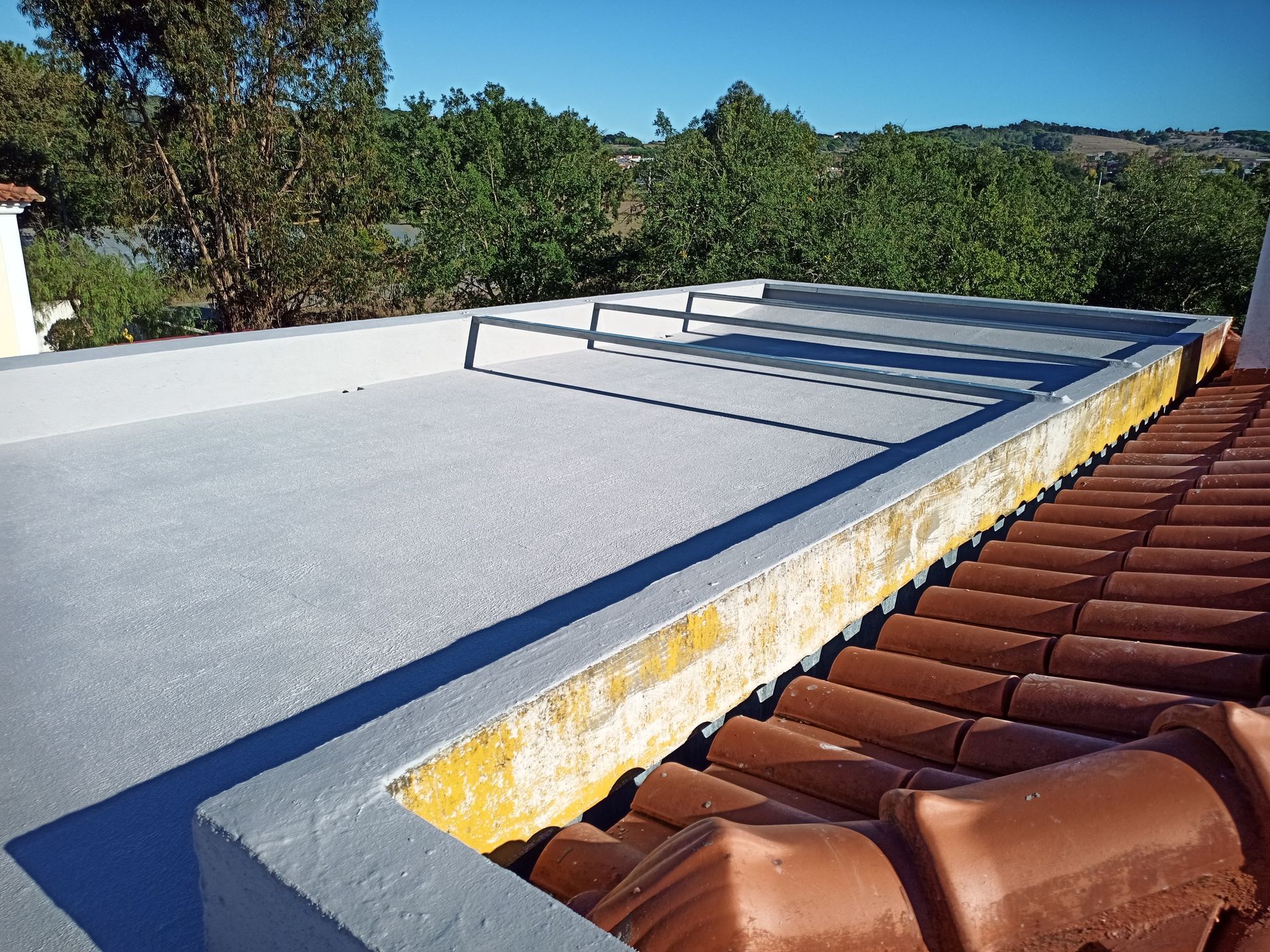 Telhado plano e cinza com parapeito, adjacente a um telhado de telhas vermelhas, e com árvores ao fundo sob um céu azul.
