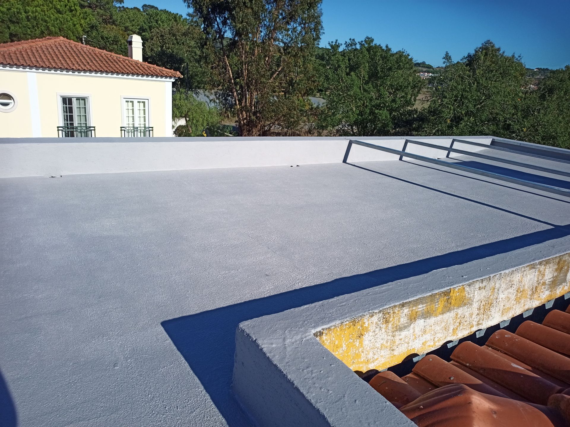 Telhado plano cinza com borda revestida de telhas, luz solar e um prédio ao fundo.