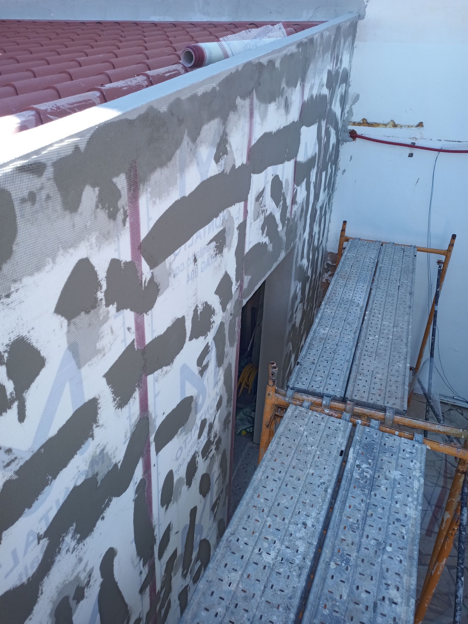 Parede externa com aplicação recente de estuque; andaimes e cobertura visíveis.