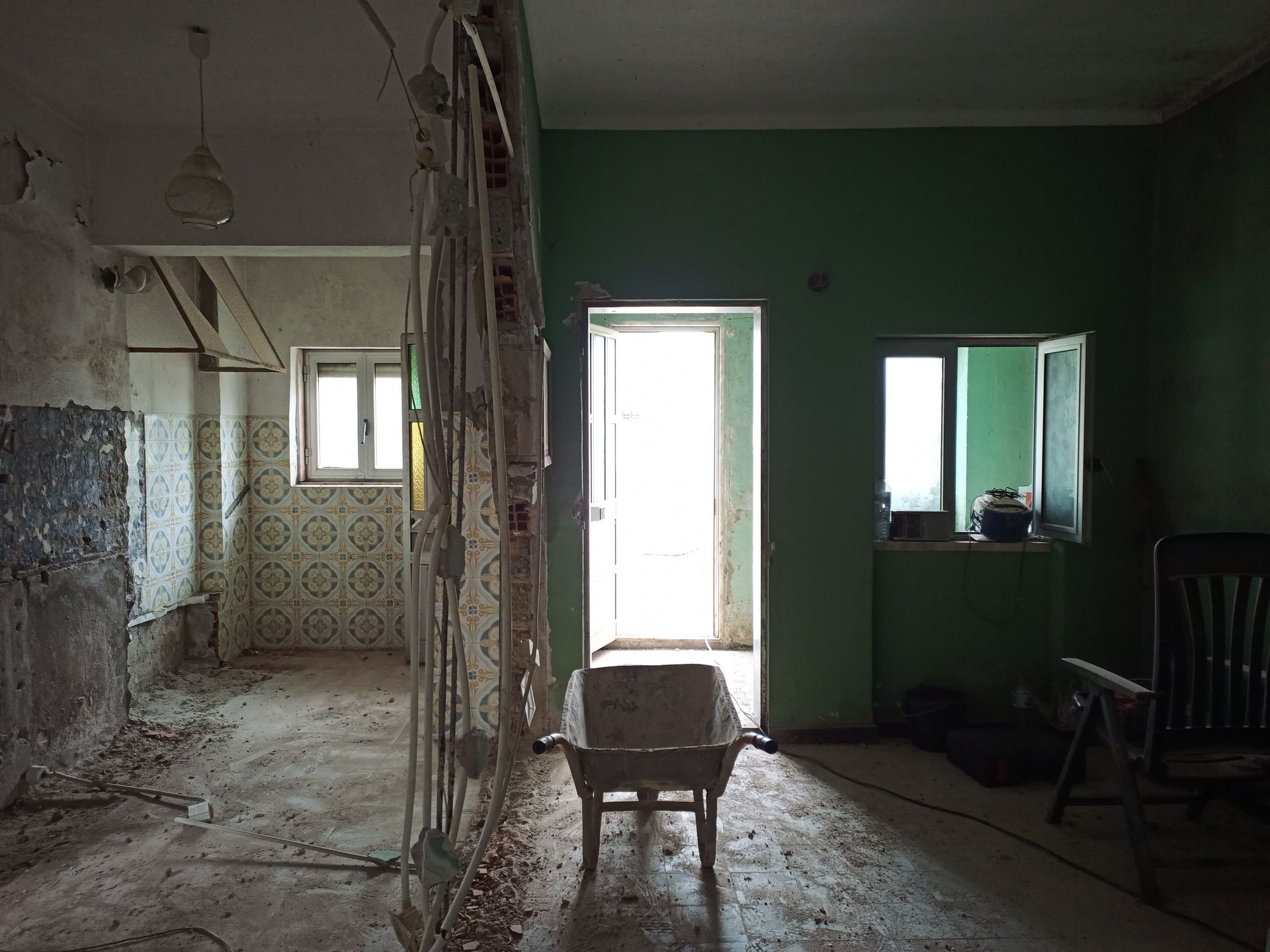 Vista interior de um cômodo em ruínas, com paredes expostas, uma porta, uma janela e uma cadeira. Parede verde visível.
