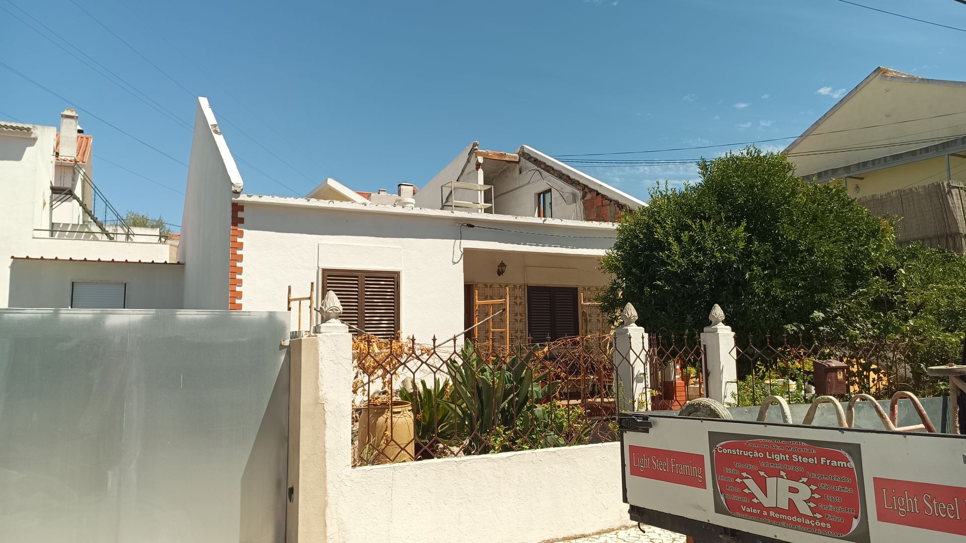 Casa branca com persianas marrons e uma árvore verdejante em frente. Céu azul brilhante.