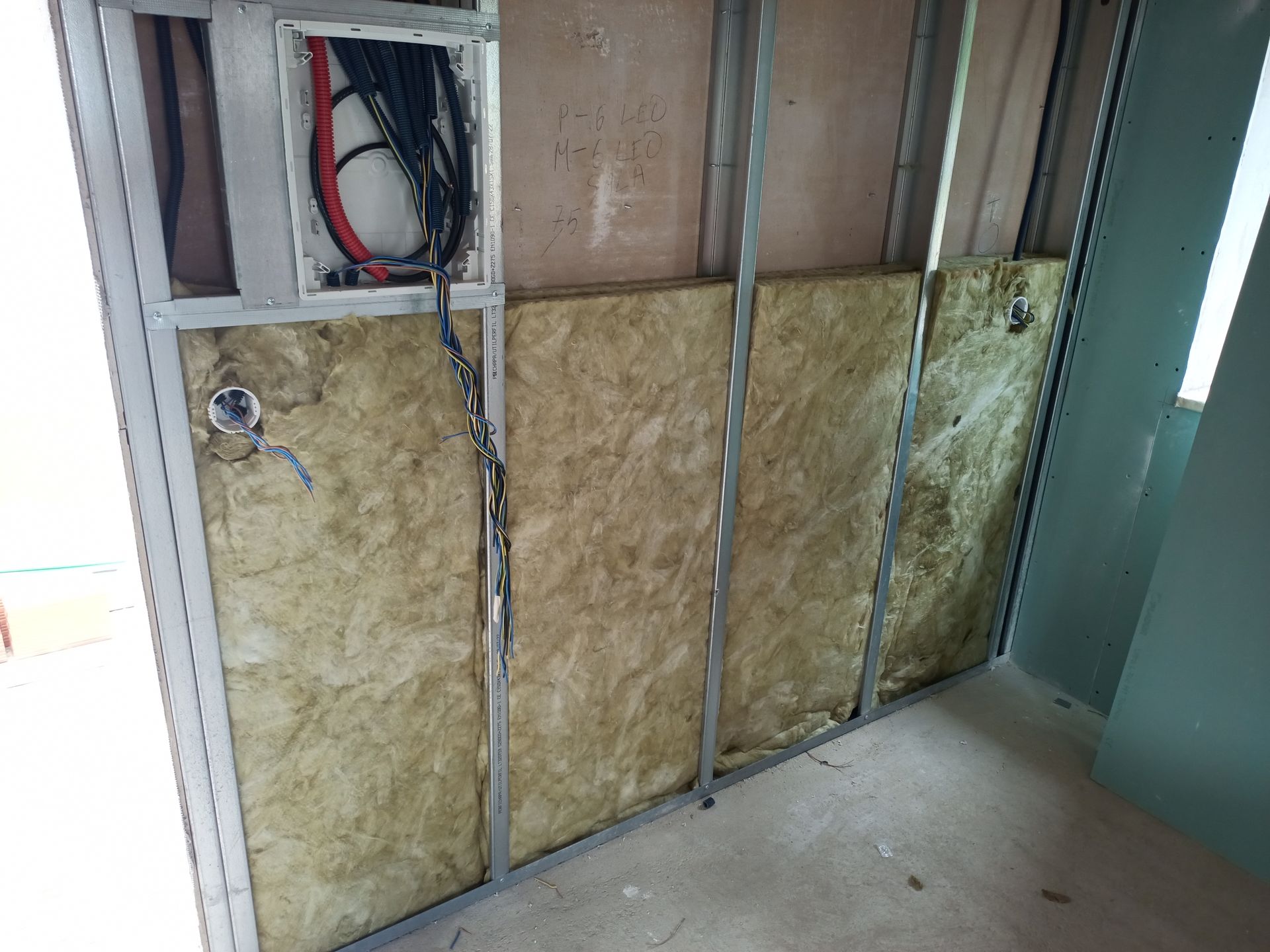 Parede interna em construção, estrutura metálica, isolamento instalado, caixa elétrica visível.