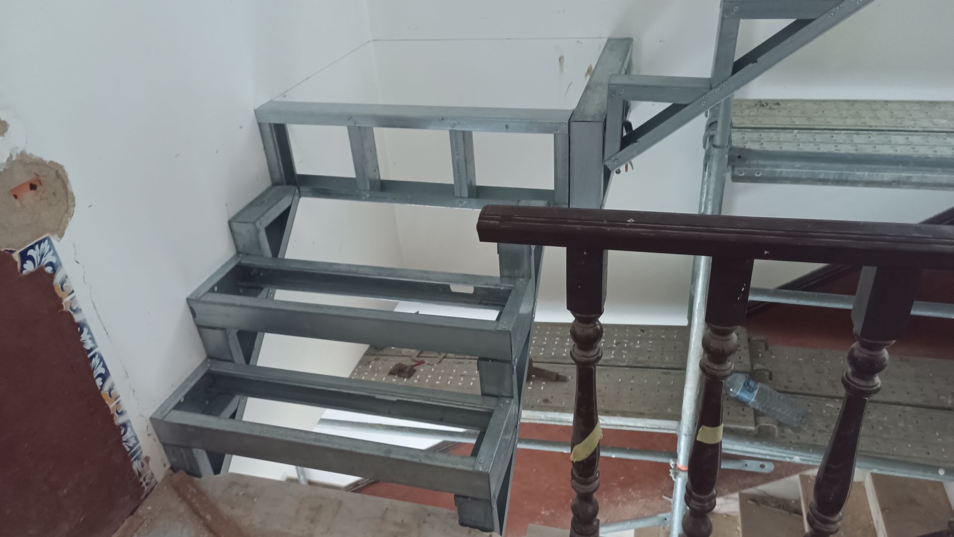 Escada com estrutura metálica. Os degraus são delineados em cinza, contrastando com uma parede branca. Possui corrimão de madeira.