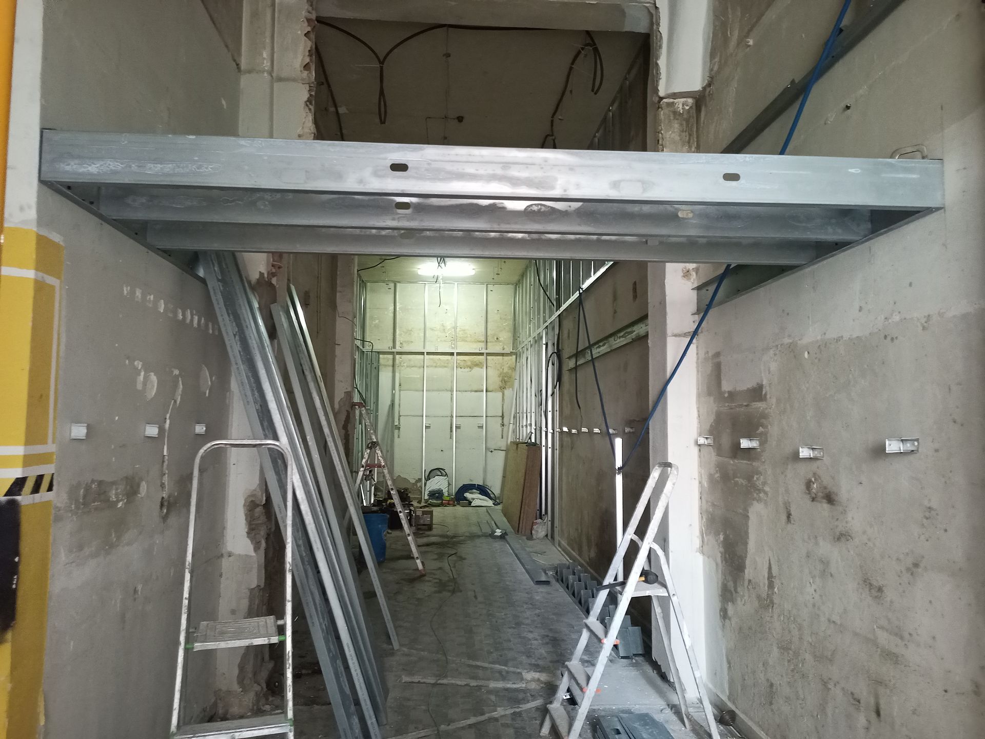 Canteiro de obras com estrutura de vigas metálicas atravessando um corredor. Escadas e materiais espalhados por toda parte.